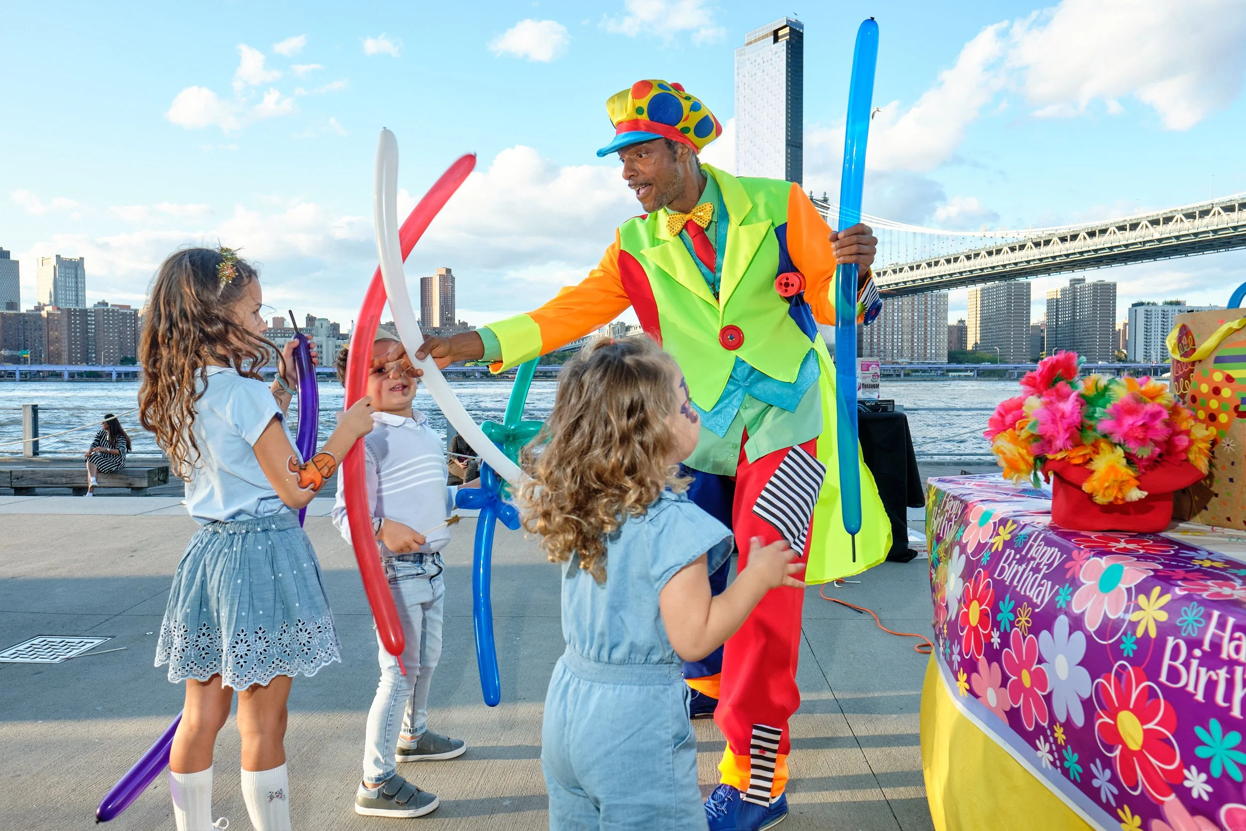 A clown in colorful costume handing out balloons to children at a birthday party outdoors with a New York Ciity skyline and bridge in the background.
