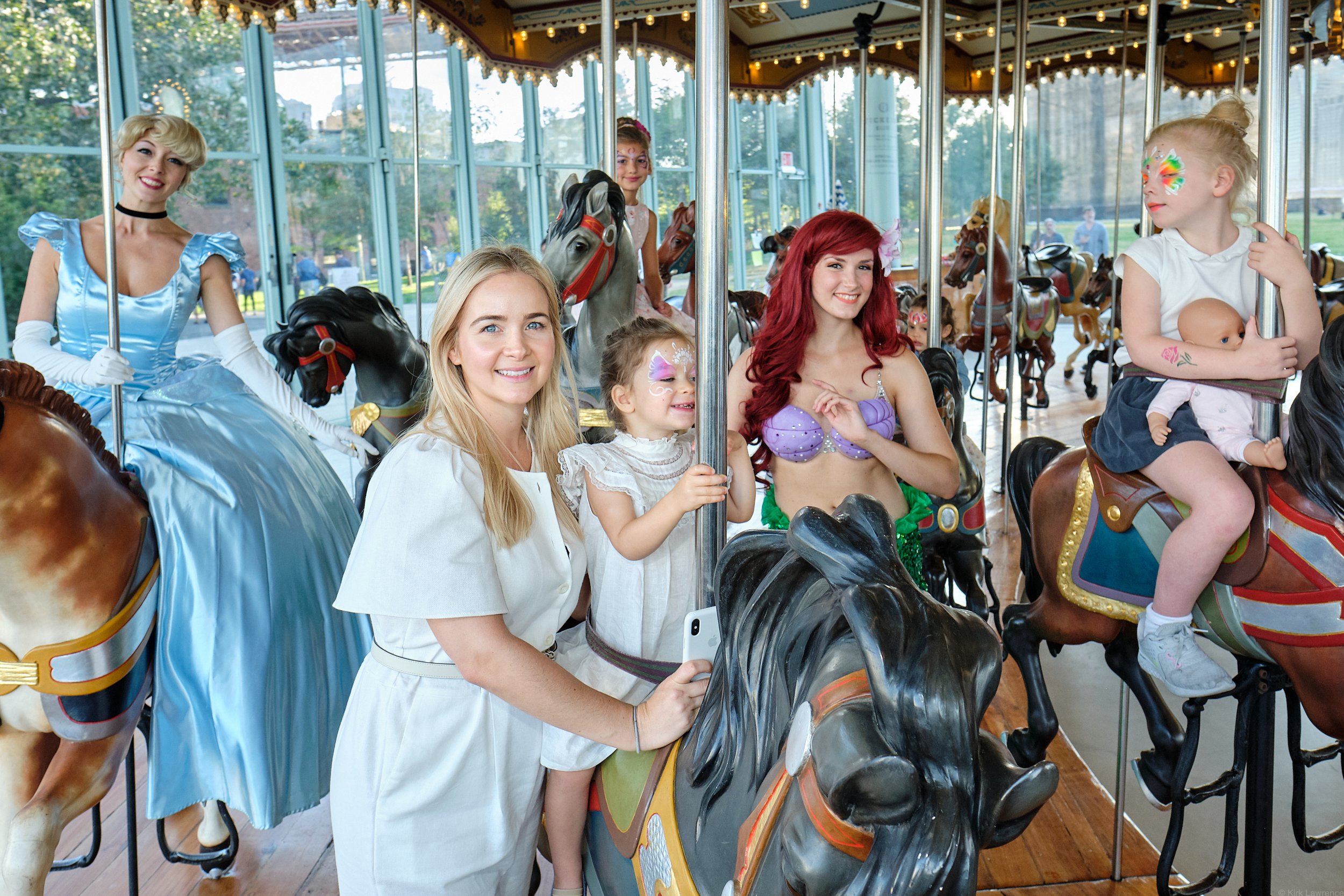 Group of women and children riding on a carousel with wooden horses and a canopy decorated with lights, inside a park with trees and people in the background.