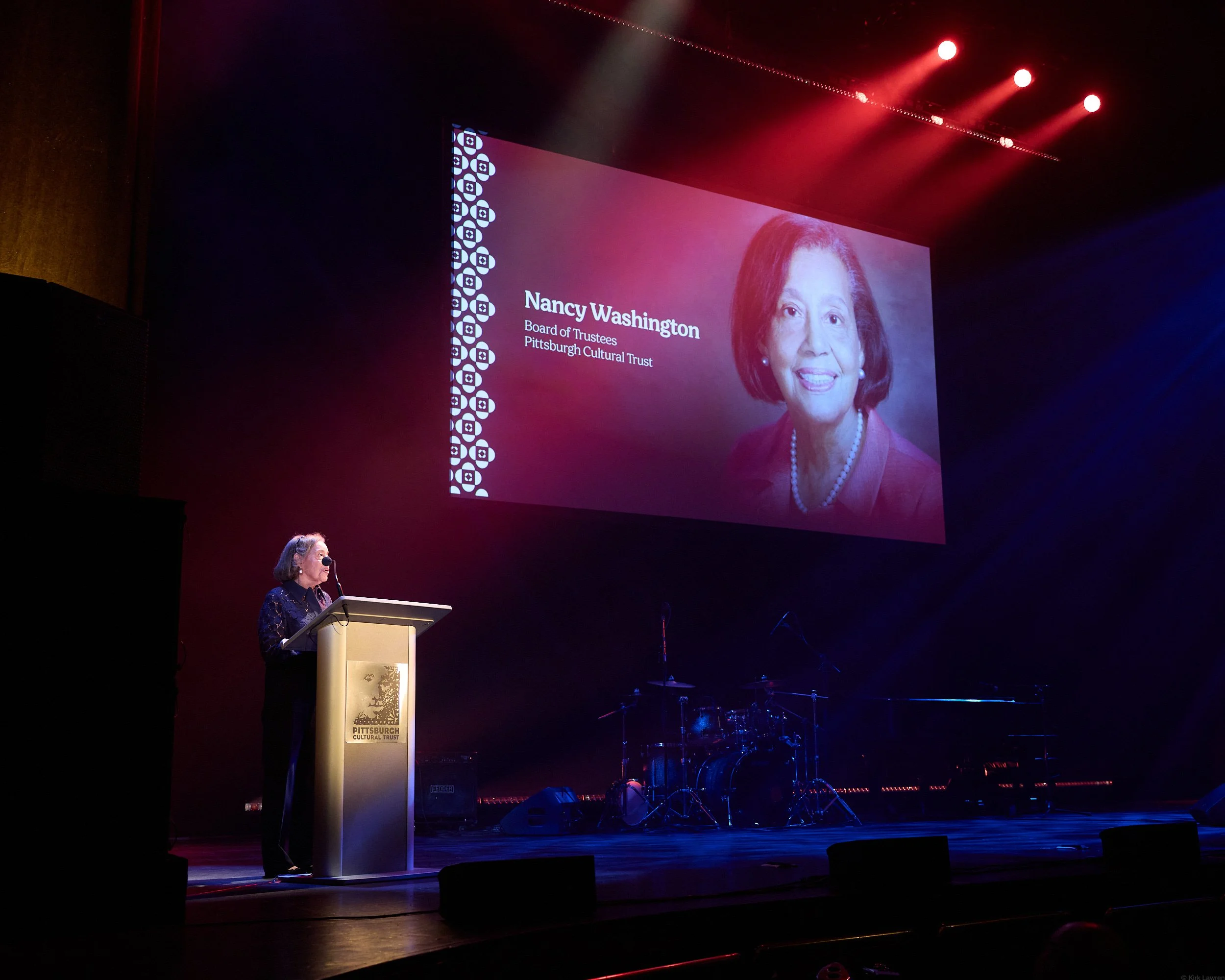 Nancy Washington speaking at a podium during a ceremony or event with a large screen displaying her name and title in the background.