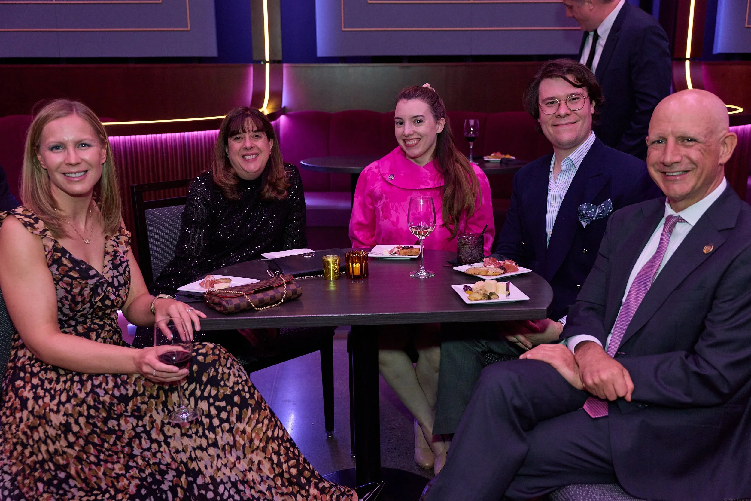 Group of six adults dressed in formal attire sitting at a table in a dimly lit restaurant or bar with purple and pink neon lighting.