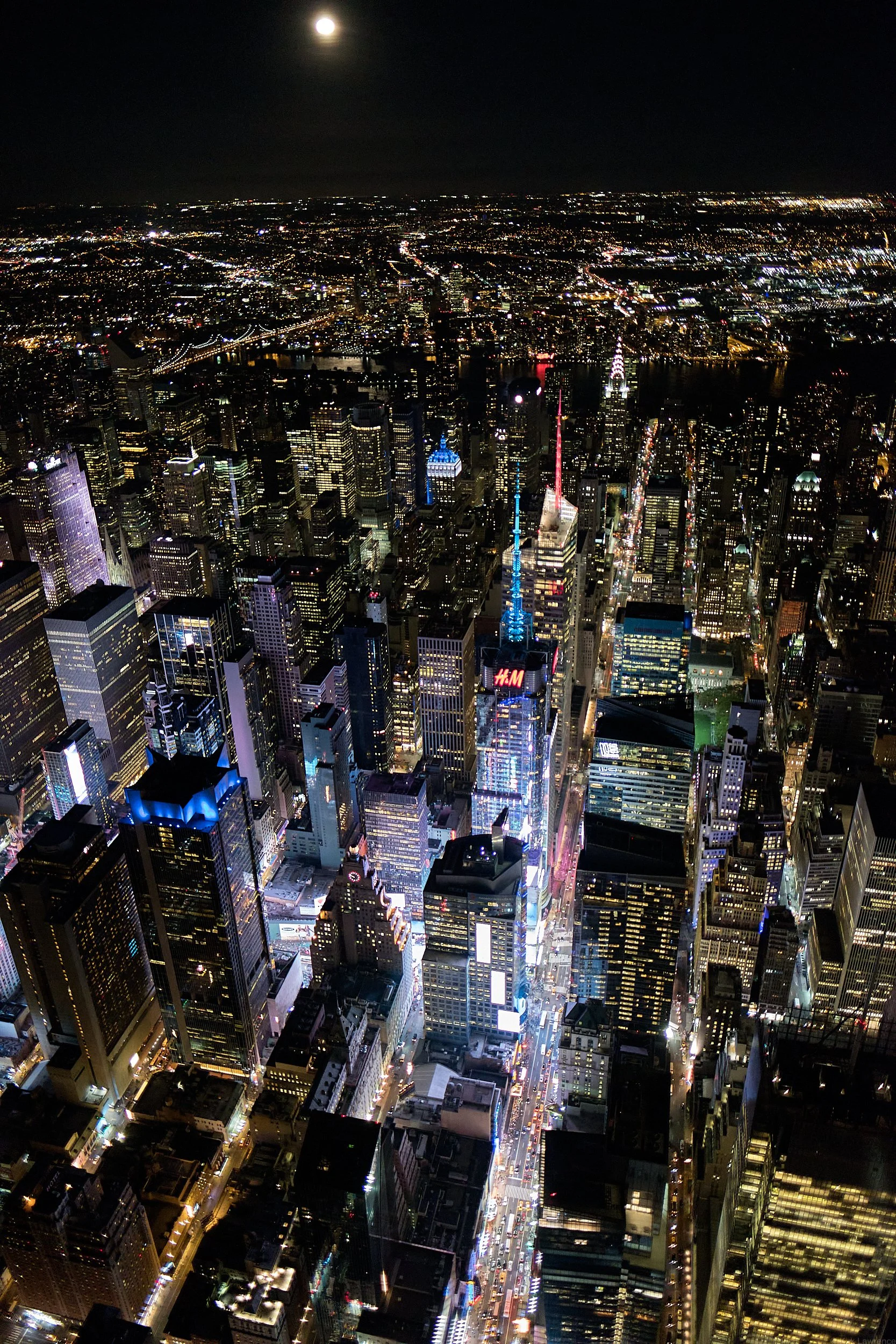 Nighttime aerial view of New York City skyline illuminated with lights, including the Empire State Building and the Chrysler Building.