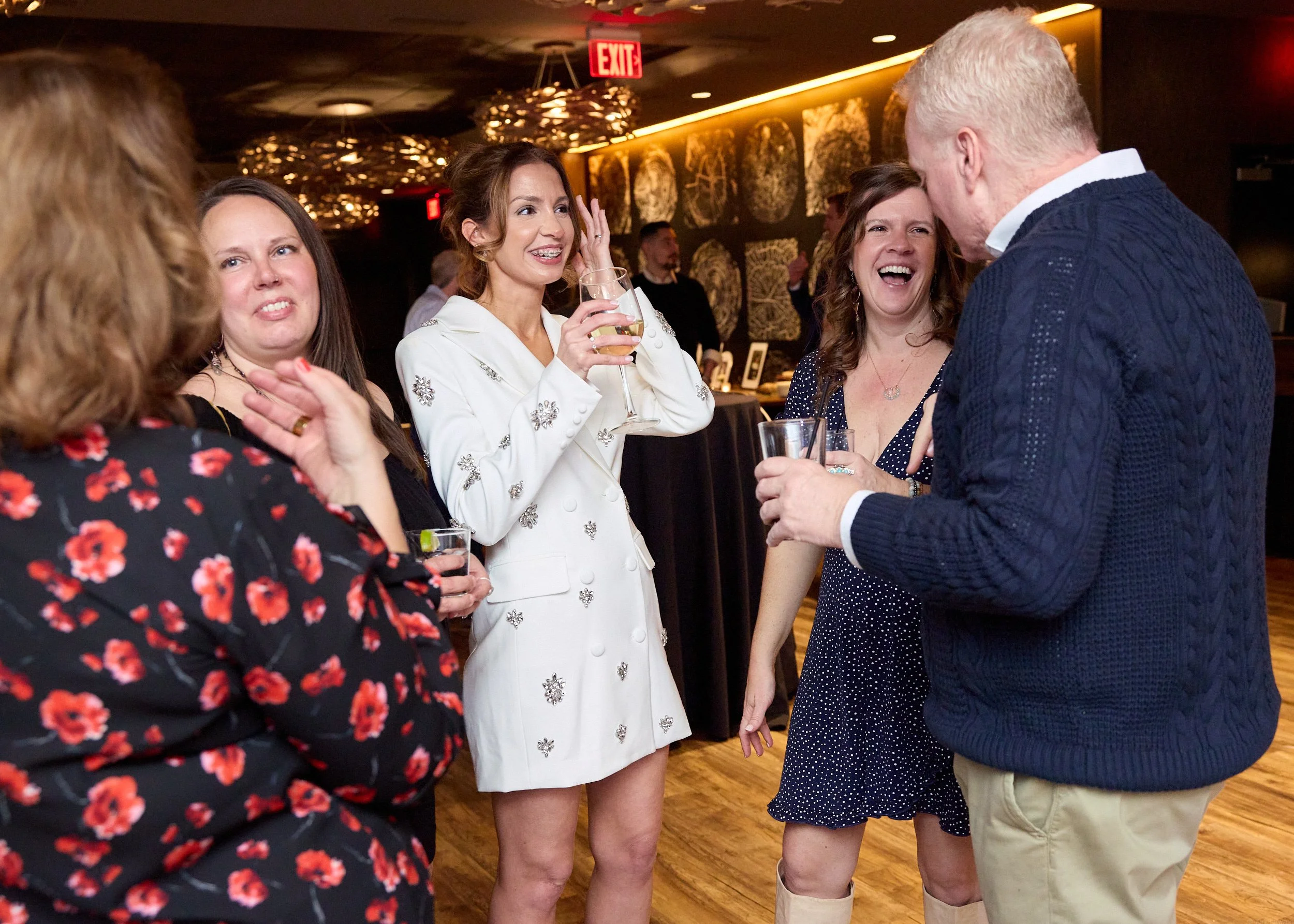 People enjoying a social gathering indoors, engaged in conversation, with drinks in hand, warmly lit decor, and wall art in the background.