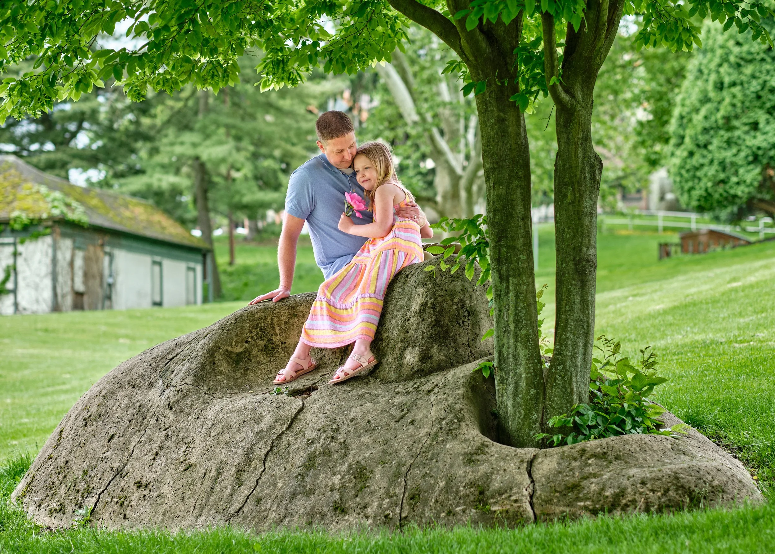 A man and a young girl sitting on a large tree root in a park, surrounded by green leaves and grass.