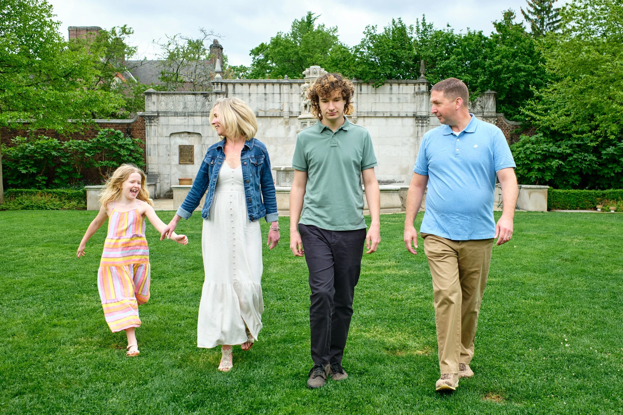 A family of five walking on a grassy lawn in front of a decorative fountain and lush green trees.