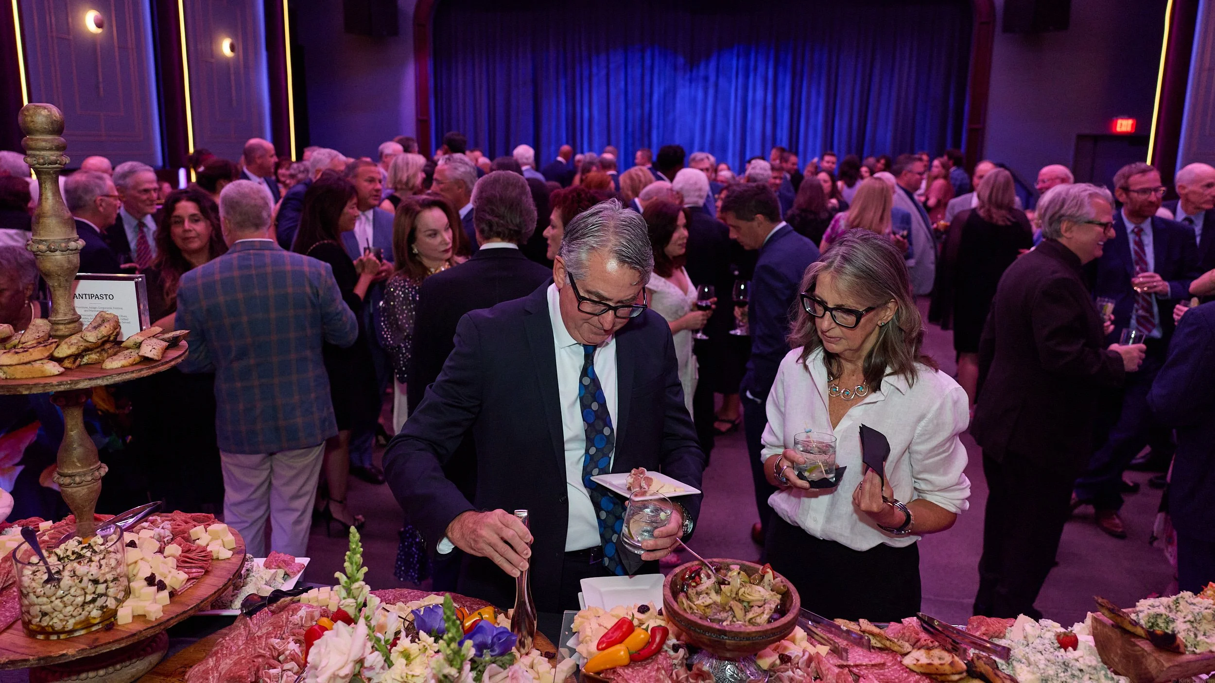 Crowd of people at a social gathering or party, with a table of food in the foreground and a blue curtain on the stage in the background.