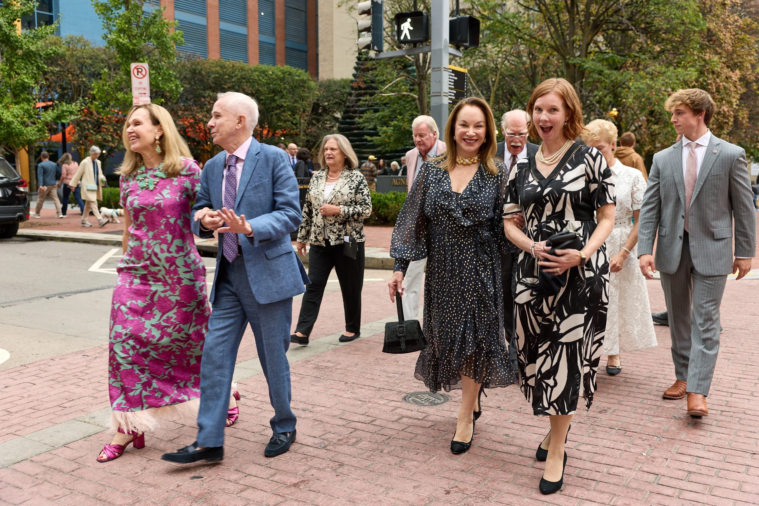 A group of mostly women and a few men dressed in formal attire walking on a city street, smiling and engaging with each other, with trees and buildings in the background.