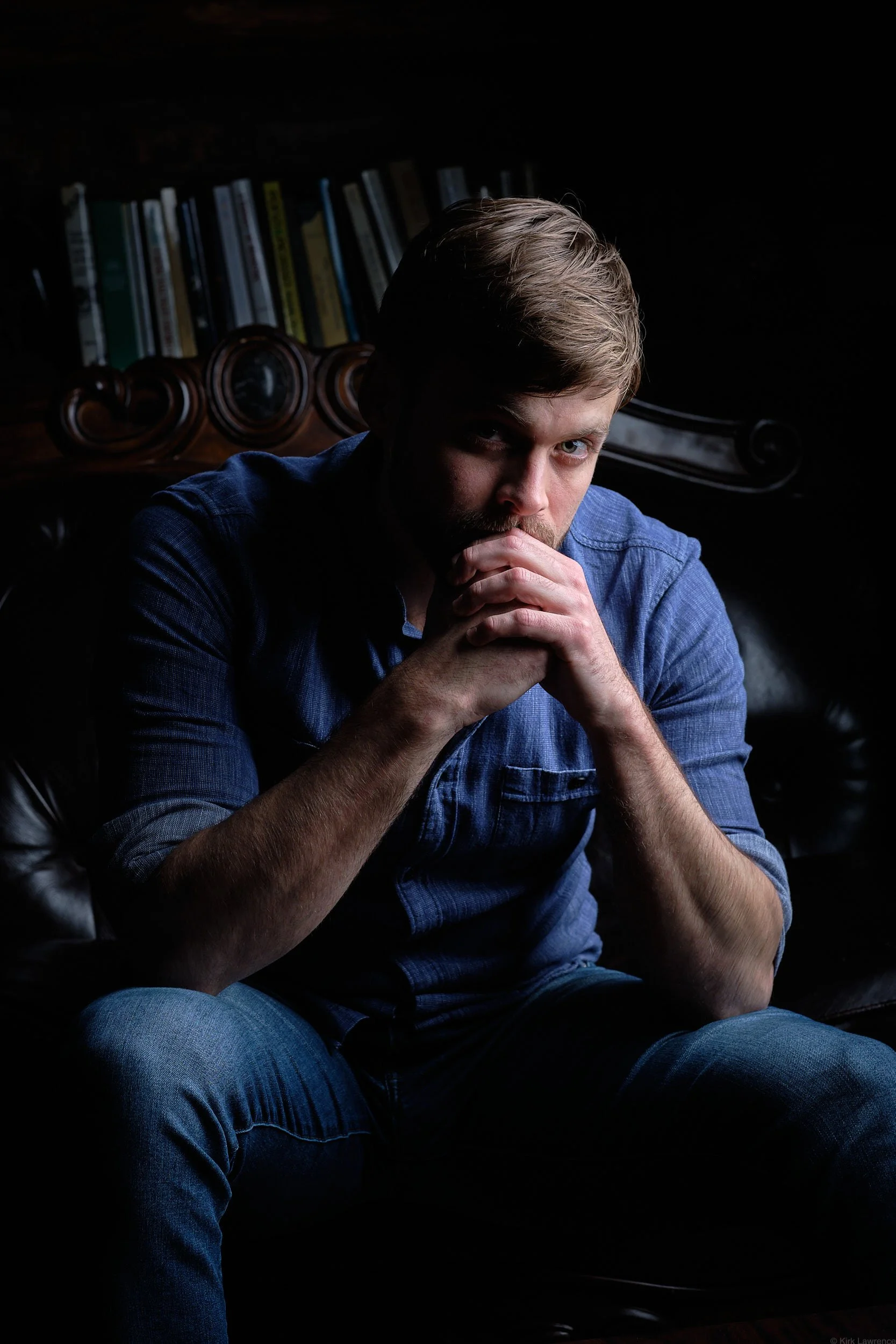 A young man with blond hair and light-colored eyes seated in a dark room, with his hands clasped near his mouth, wearing a blue shirt and jeans, sitting on a black leather chair with a wooden backrest and books in the background.