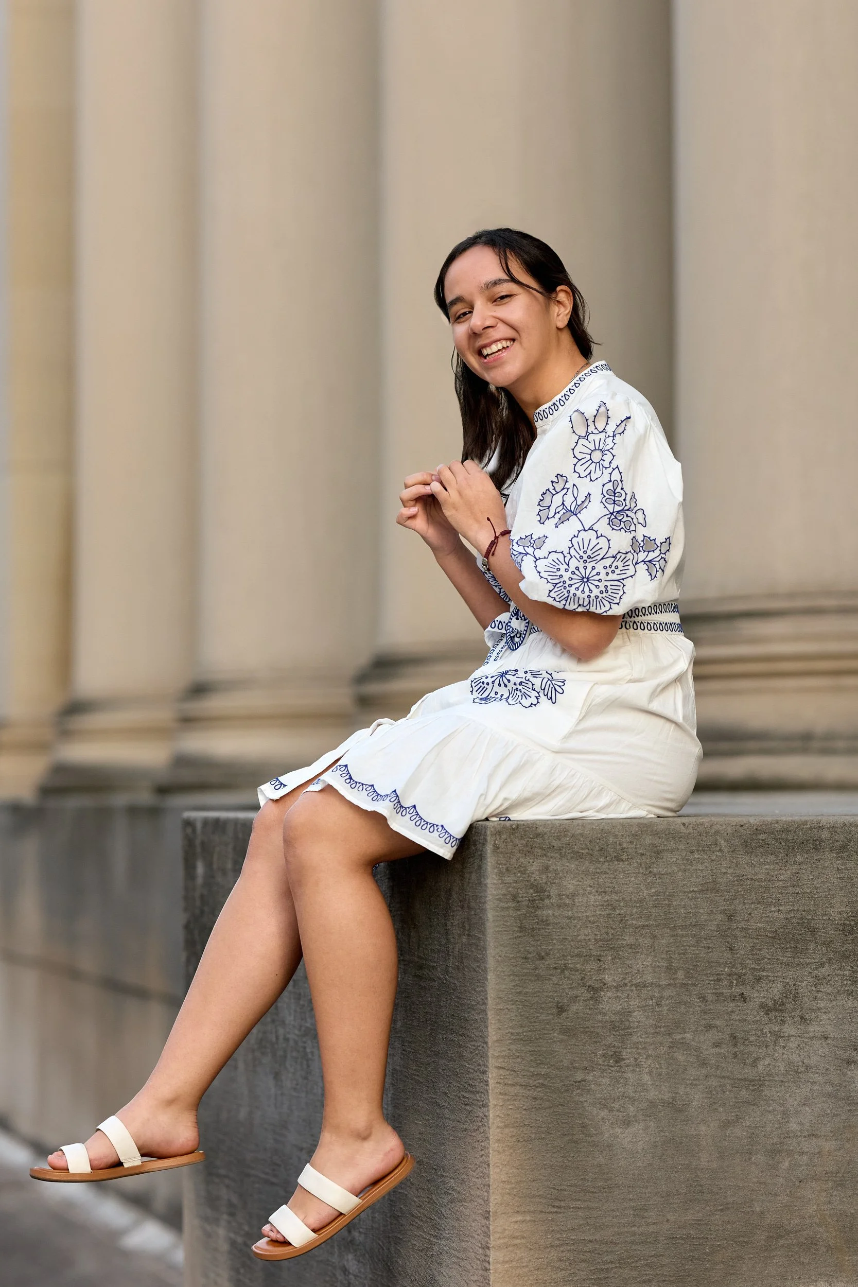 A woman sitting on a concrete ledge with her feet dangling, wearing a white dress with blue embroidery and white sandals, smiling.