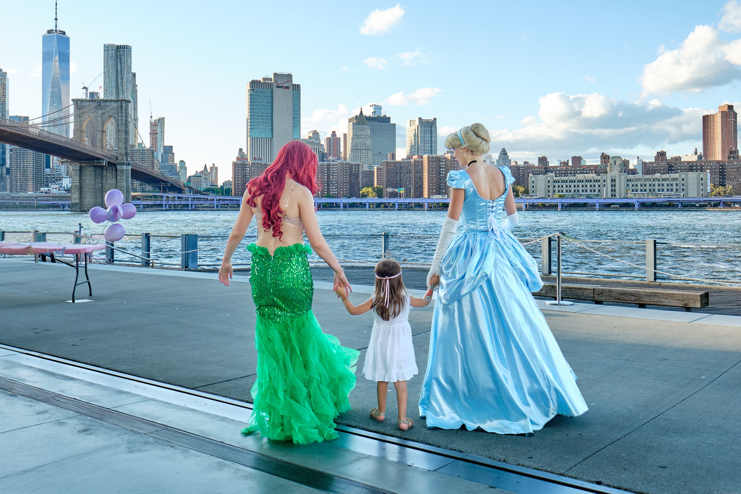 Two women dressed as Disney princesses, Ariel and Cinderella, holding hands with a young girl in a white dress by the water in New York City with the Brooklyn Bridge and Manhattan skyline in the background.