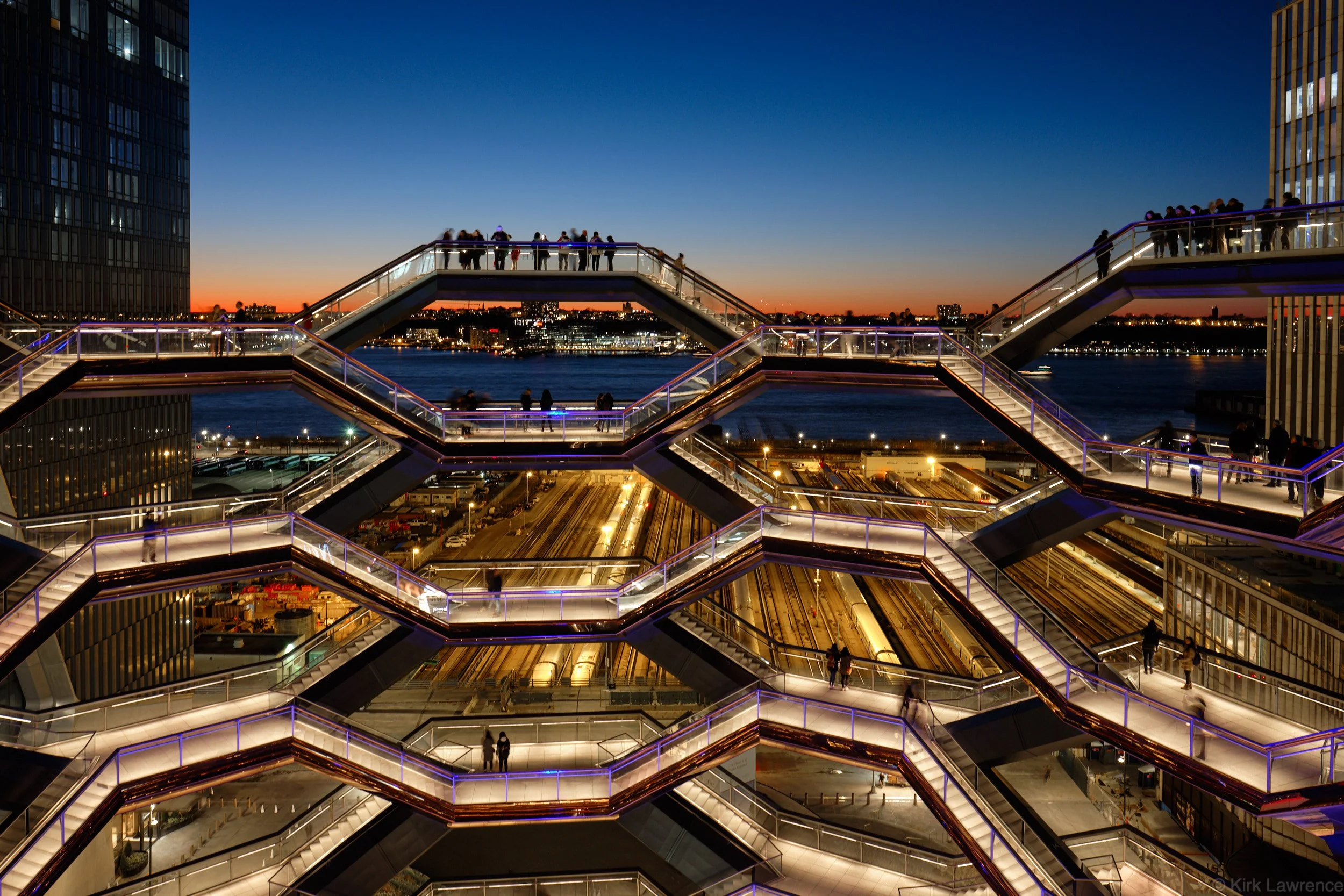View of a high-rise building's skybridge with illuminated stairs at dusk, overlooking a New York City cityscape with river and sunset sky in the background.