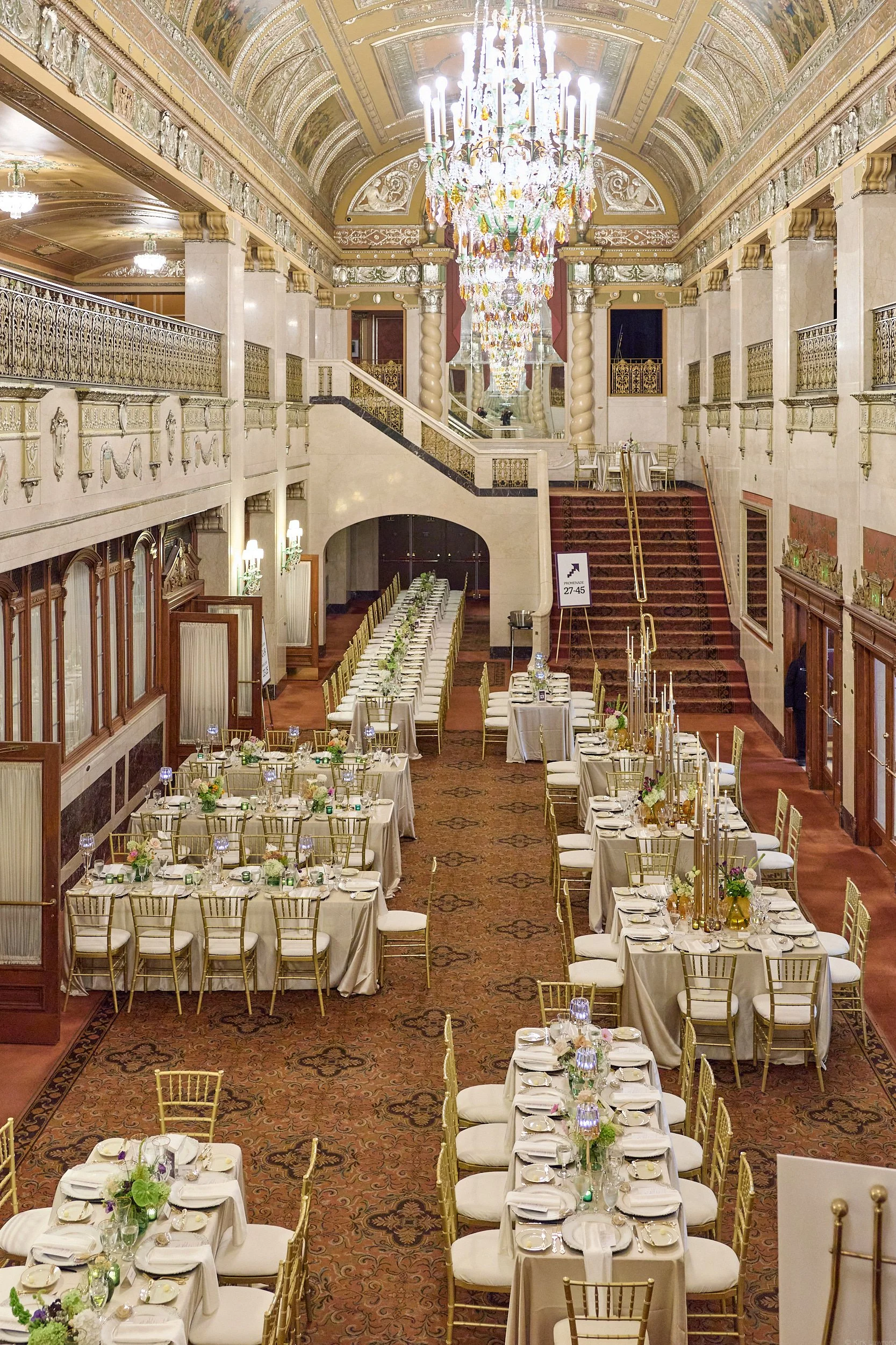 Elegant banquet hall with long tables set with white tablecloths, gold chairs, and floral centerpieces. Large chandelier hangs from ornately decorated ceiling, with gilded railings and staircase in the background.
