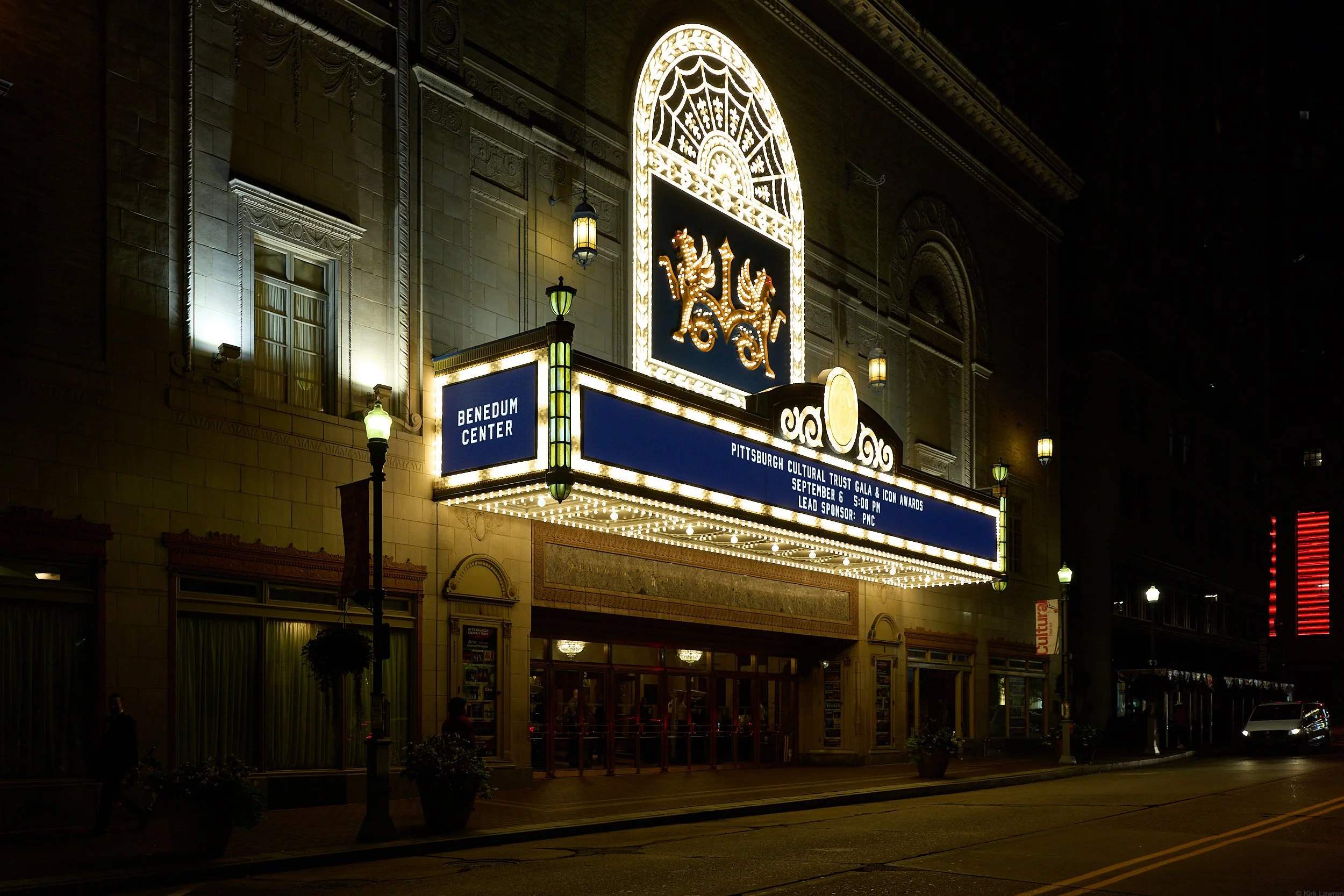 Nighttime view of the Benedum Center, a historic theater in Pittsburgh, with illuminated signs and decorative lights on its facade. The marquee displays text about an event hosted by the Pittsburgh Cultural Trust.