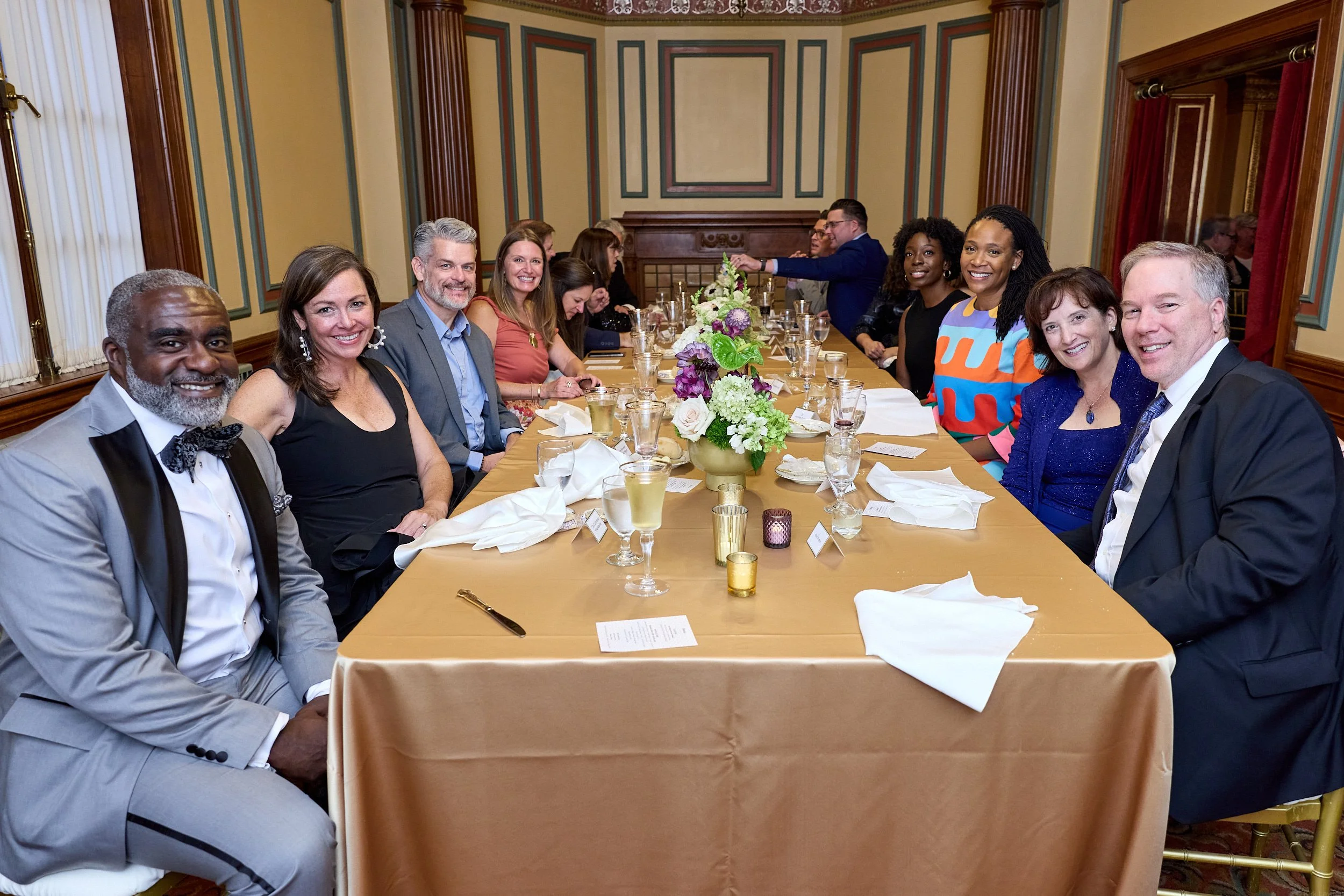 People sitting at a banquet table in a formal room with wood paneling, colorful wall decor, and a fireplace, dressed in formal attire, smiling at the camera.