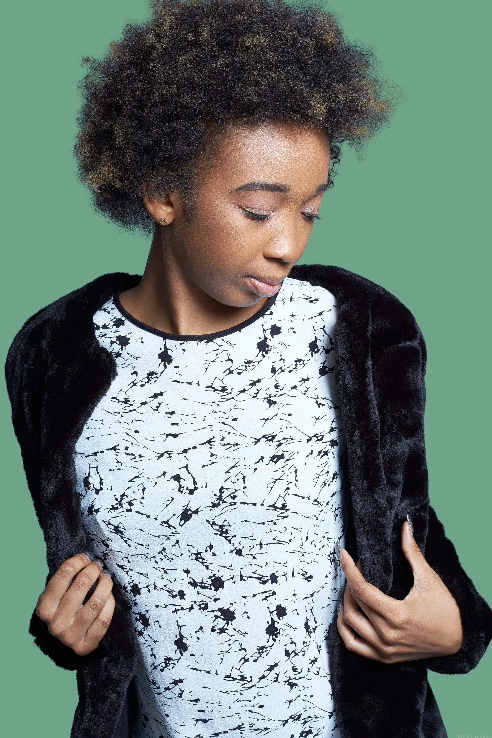 A woman with curly, mixed-tone hair in a natural style, wearing a black and white patterned top and a black jacket with a furry texture, standing against a green background.