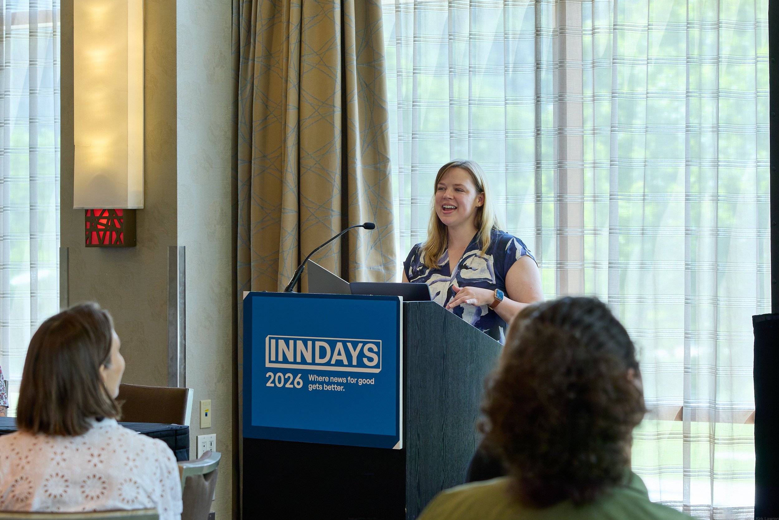 A woman speaking at a conference in front of an audience, standing behind a podium with a sign that reads "INN DAYS 2026 Where news for good gets better," in a room with large windows and curtains in the background.