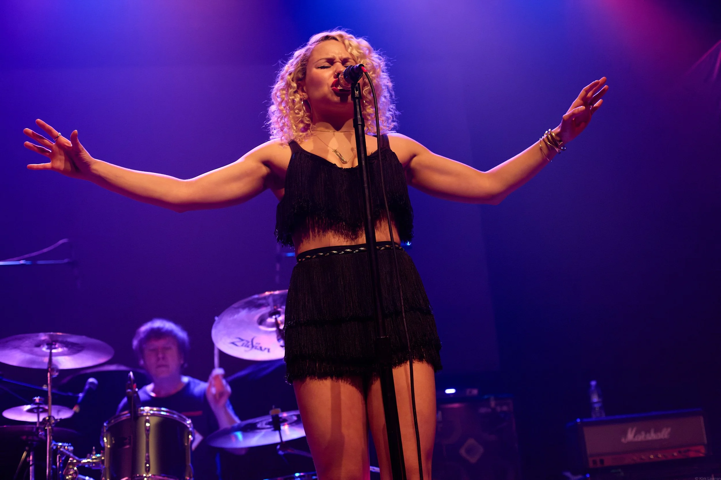 A woman with curly blonde hair singing into a microphone on stage with purple lighting, wearing a black fringed dress and jewelry, with a drummer in the background.