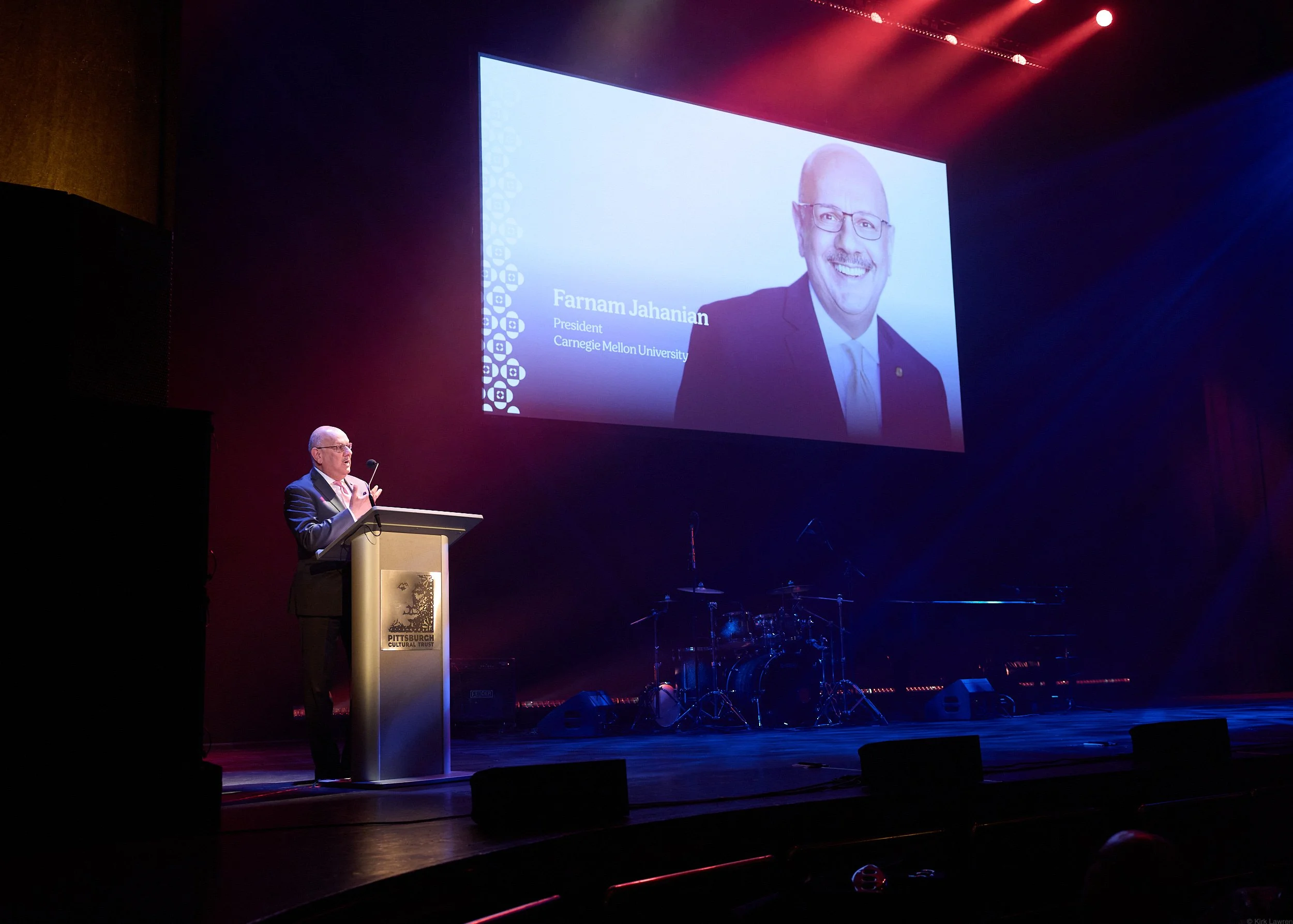 A man is speaking at a podium at an event, with a large screen behind him displaying his photo, name (Farnam Jahanian), his title (President), and affiliation (Carnegie Mellon University). The stage is dimly lit with colorful lights, and musical inst