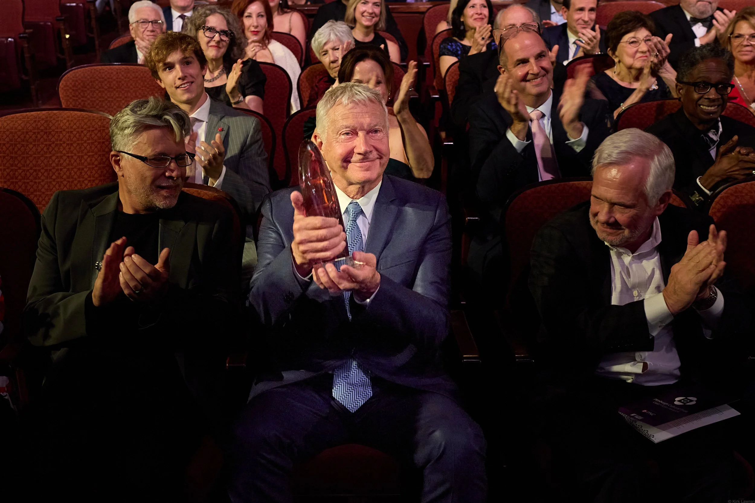 An elderly man in a suit holding a trophy and smiling at a formal event surrounded by laughing and clapping audience members in a theater with red seats.