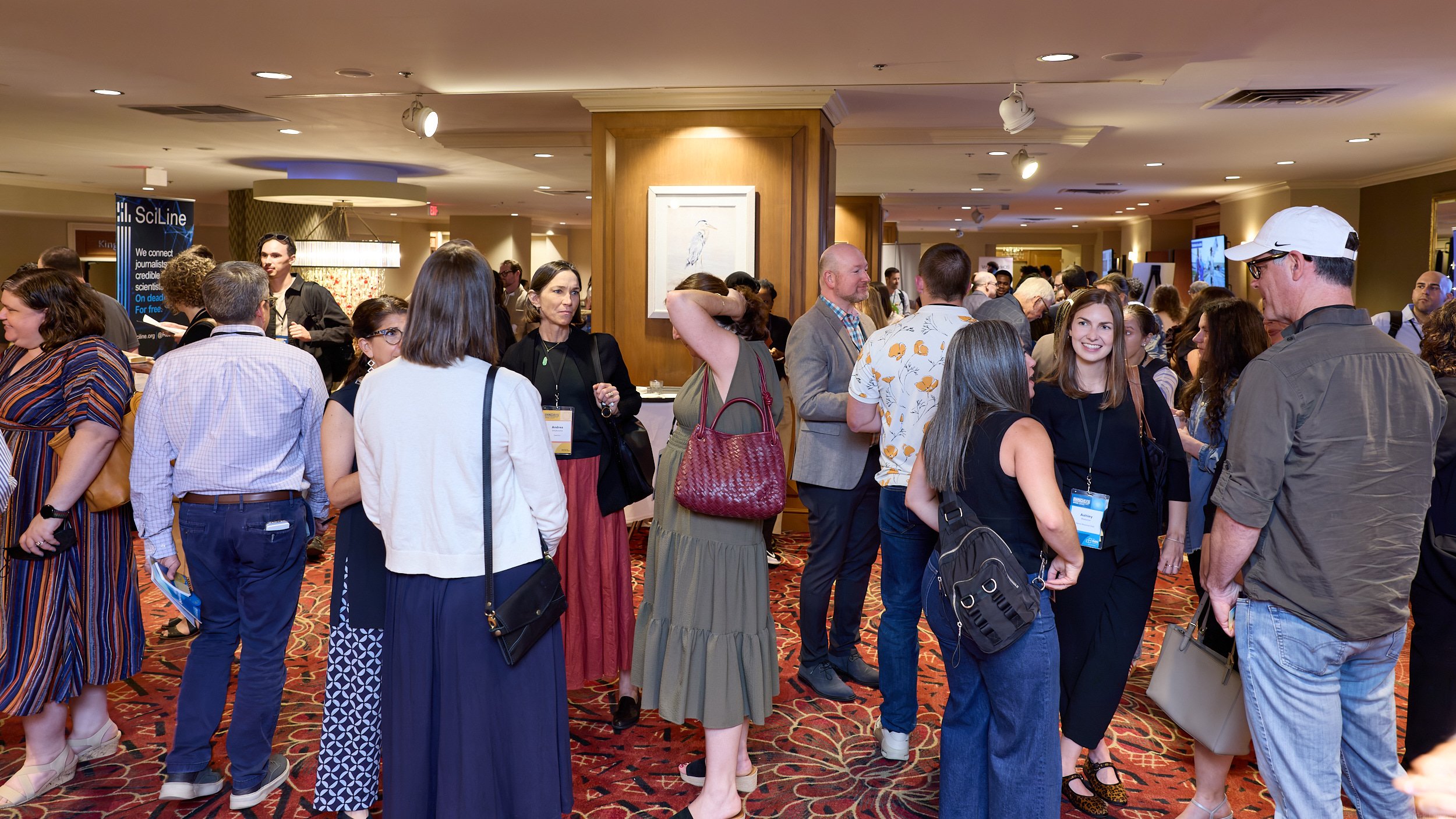 A large group of people gathered in a hotel conference lobby, engaging in conversations, with colorful carpet, wooden wall panels, and a banner in the background.