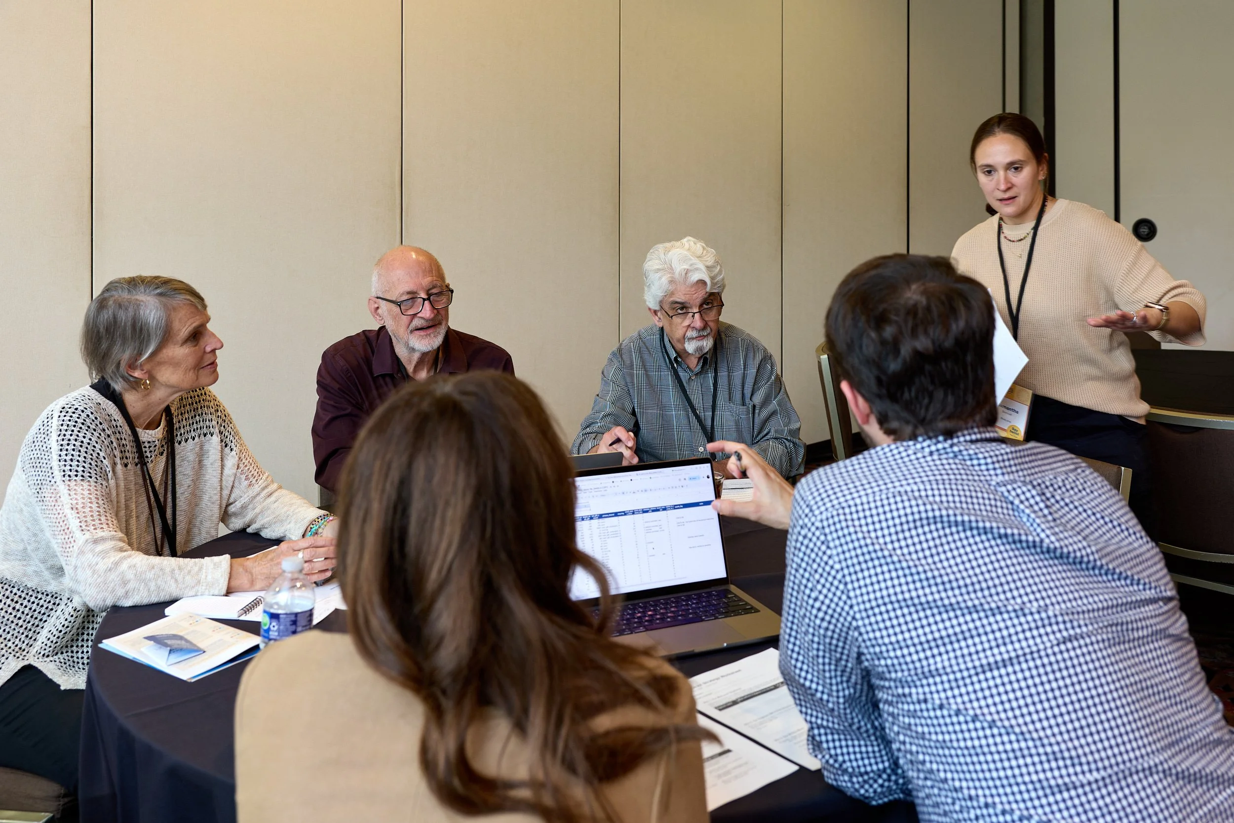 A group of people sitting around a table during a business meeting or workshop, with a woman standing and presenting in a conference room.