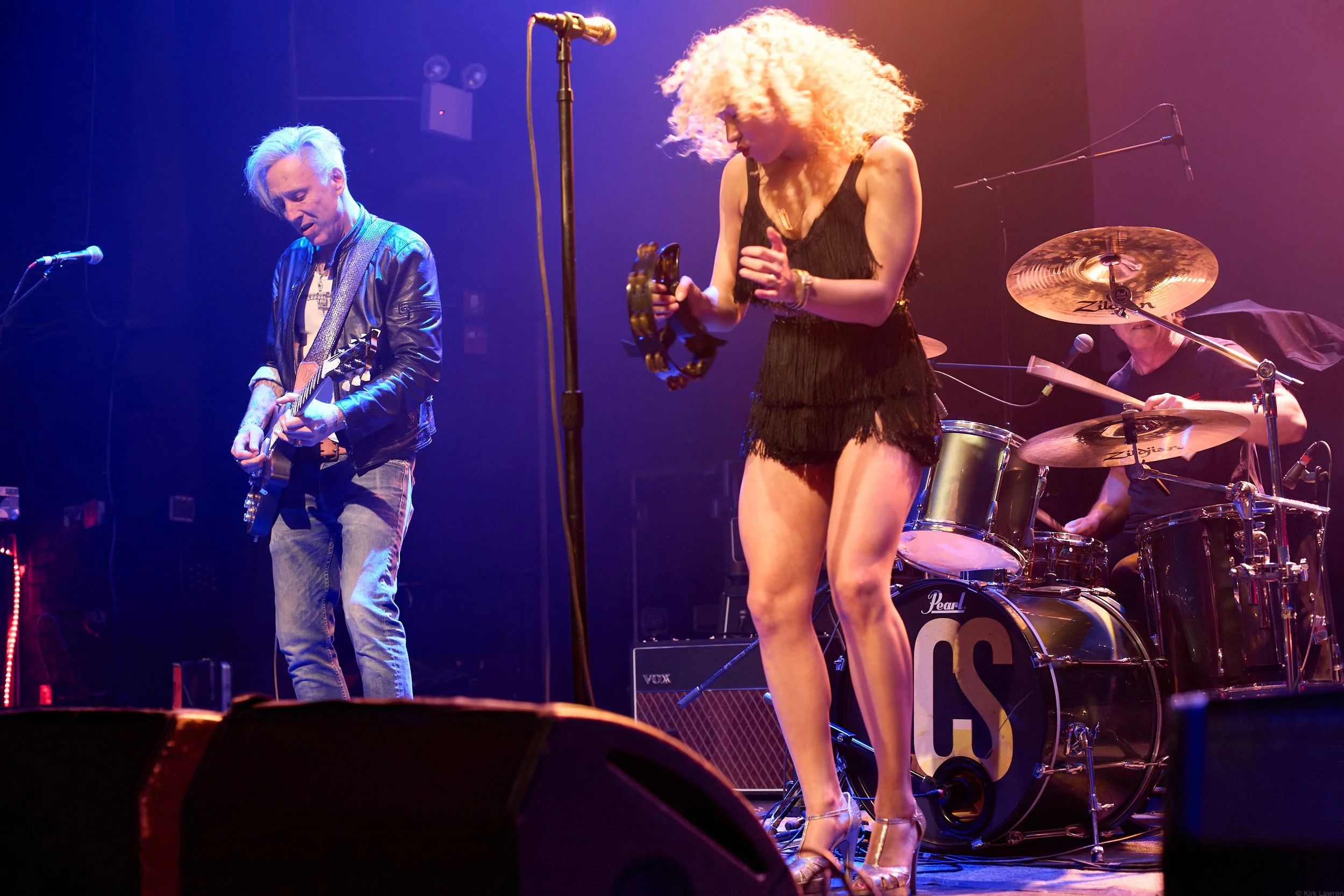 A band performing on stage with a guitarist, a female singer, and a drummer. The guitarist is wearing a leather jacket, the singer has curly blonde hair and is holding a tambourine, and the drummer is partially visible behind a drum kit. The stage is