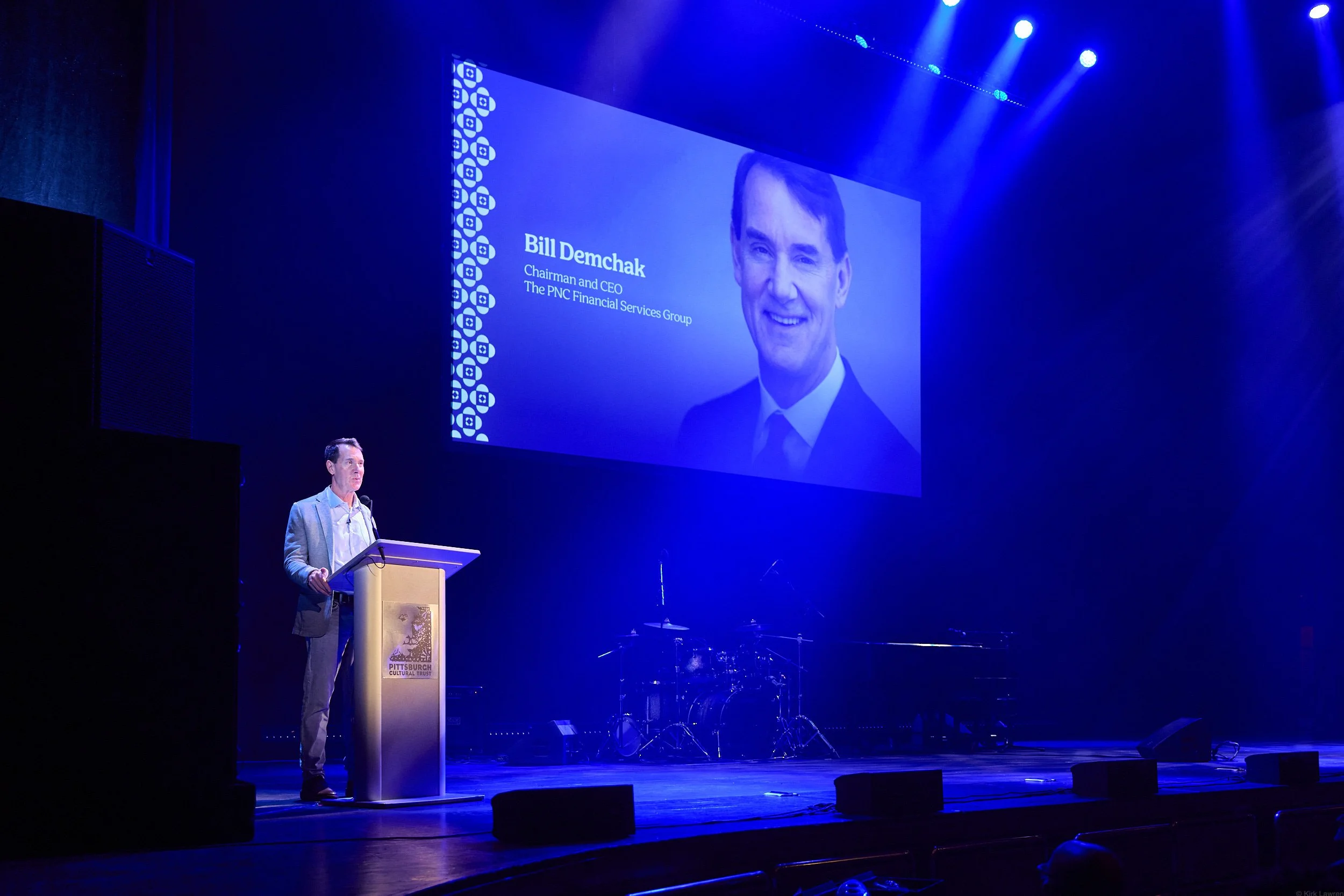 A man stands at a podium on a stage, delivering a speech during an event. Behind him, a large screen displays a portrait of another man with the name Bill Demchak and his titles, Chairman and CEO of The PNC Financial Services Group. The stage is illu