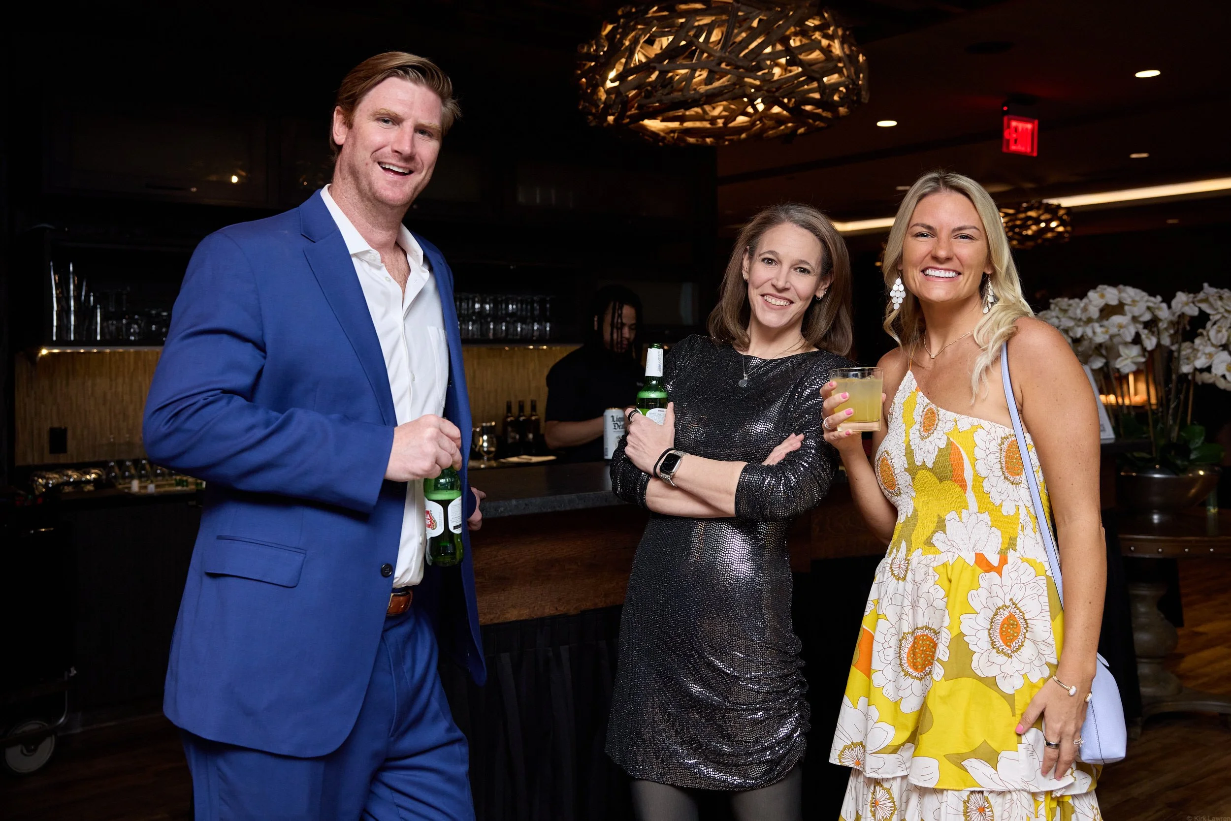 Three people at a social event, smiling and holding drinks, standing in a dimly lit venue with decorative lighting and flowers in the background.