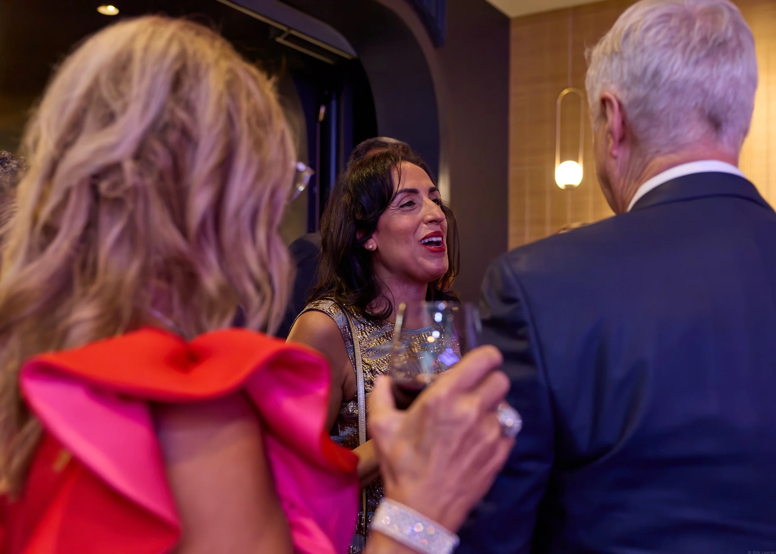 A woman with dark hair, wearing a sparkle dress, smiling, talking to a man with white hair, wearing a tuxedo, at a social event or party. A woman with blonde hair and a red dress is partially visible in the foreground.