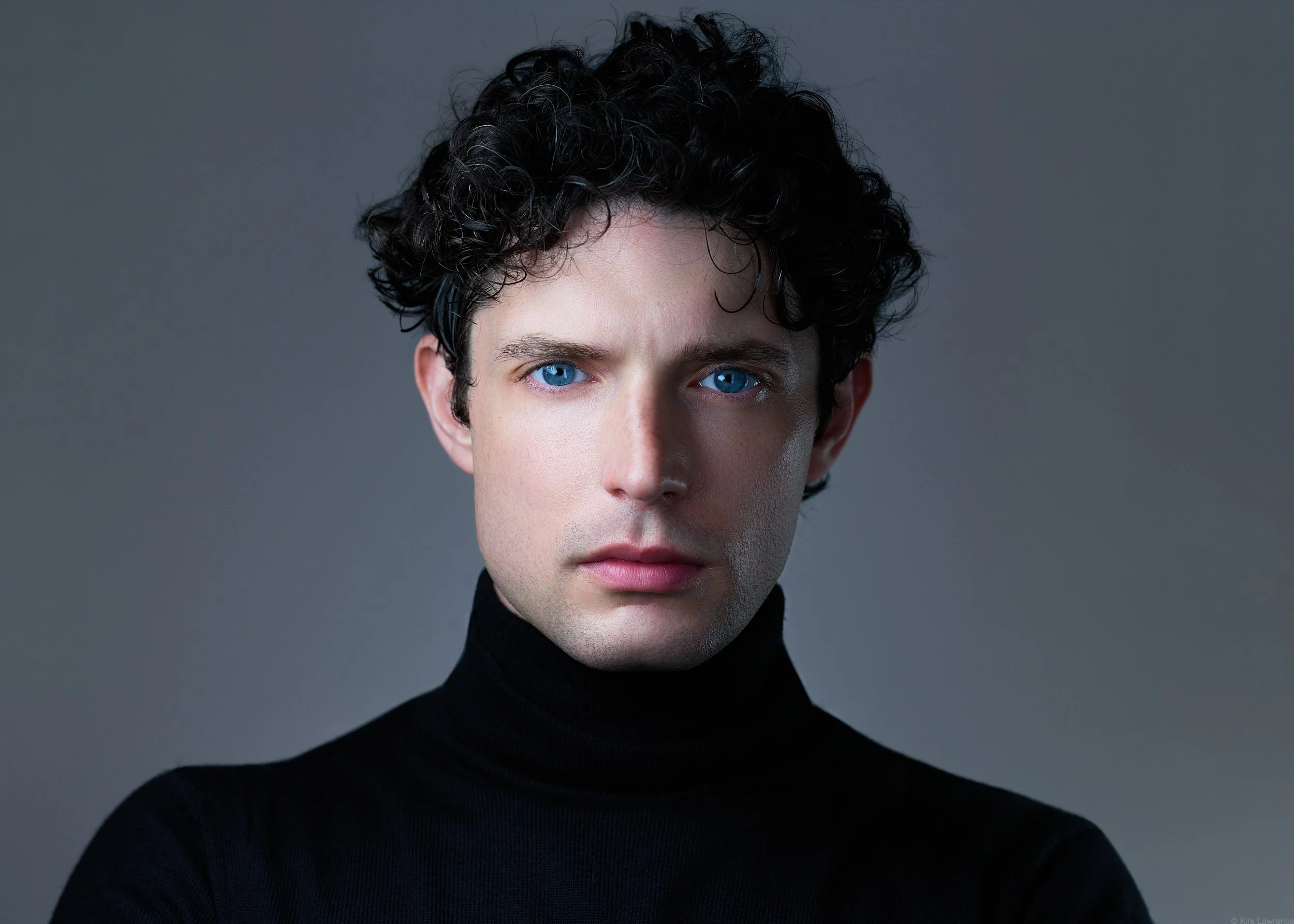 A close-up portrait of a person with curly black hair, striking blue eyes, and fair skin, wearing a black turtleneck sweater against a plain gray background.