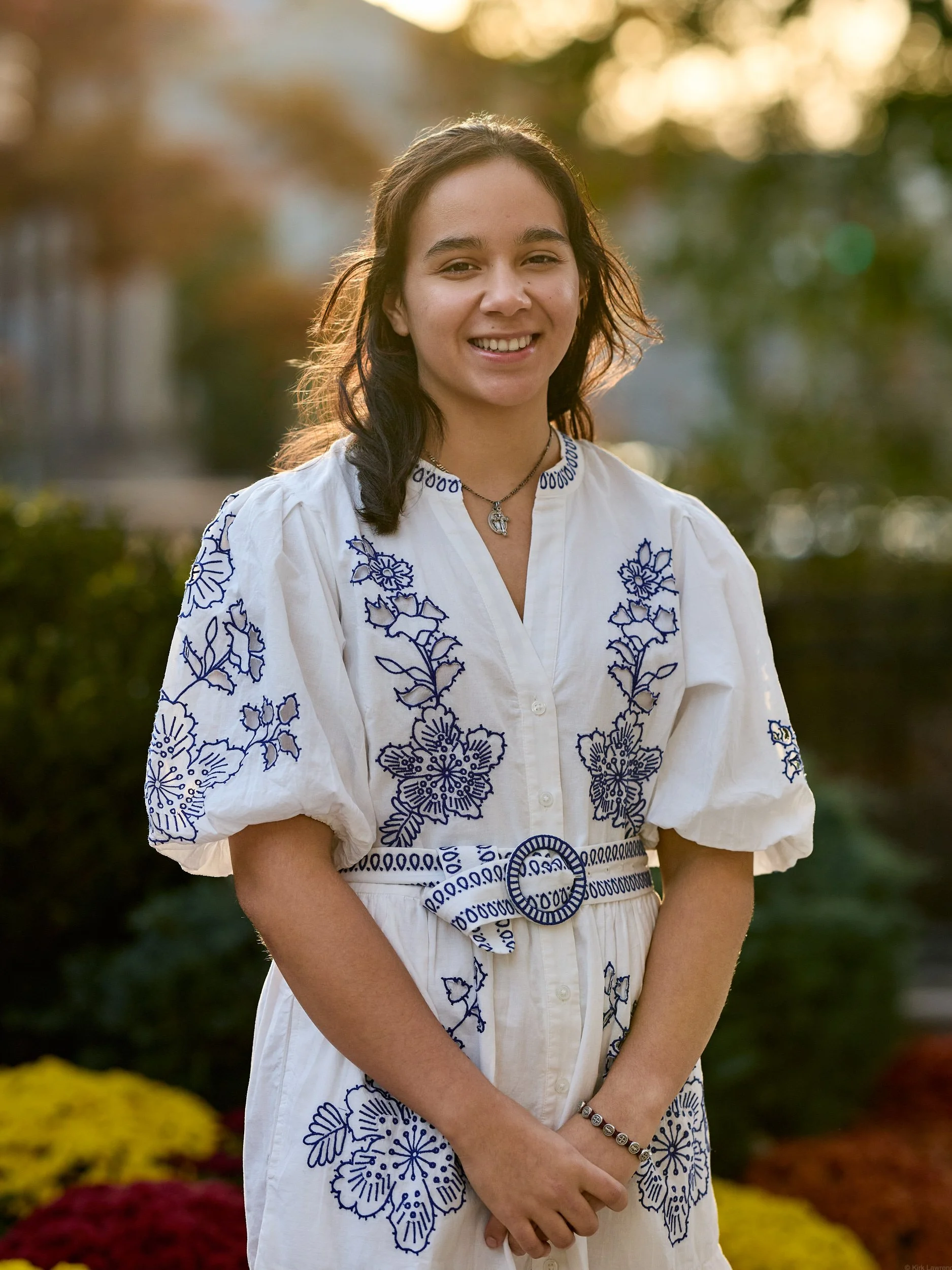 A young woman smiling outdoors in front of a background with autumn-colored trees. She is wearing a white embroidered dress with blue floral patterns, a necklace, and a bracelet, standing with her hands clasped.
