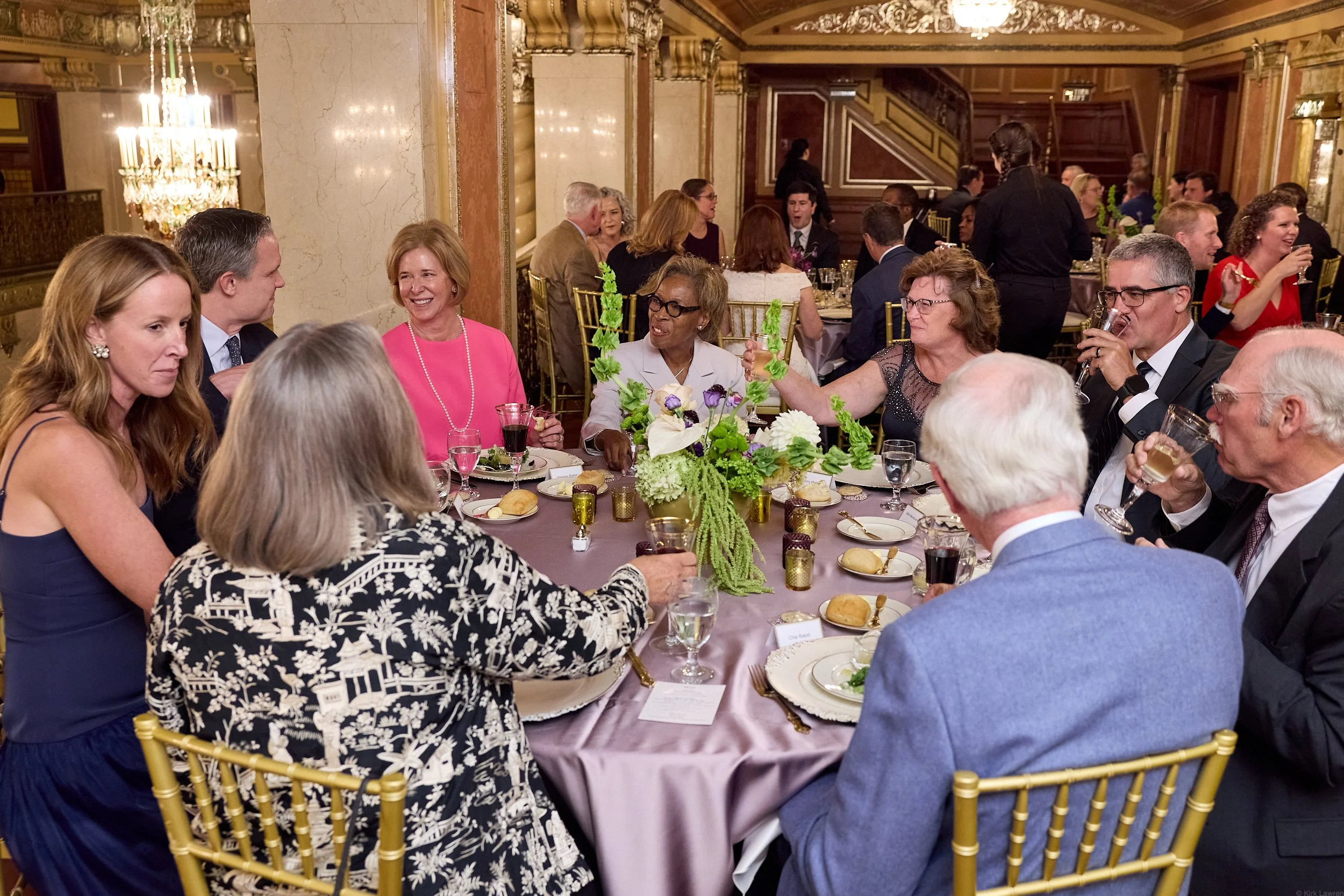 Elegant dinner event with guests seated around a table, floral centerpieces, and a woman in a pink dress smiling and talking to others.