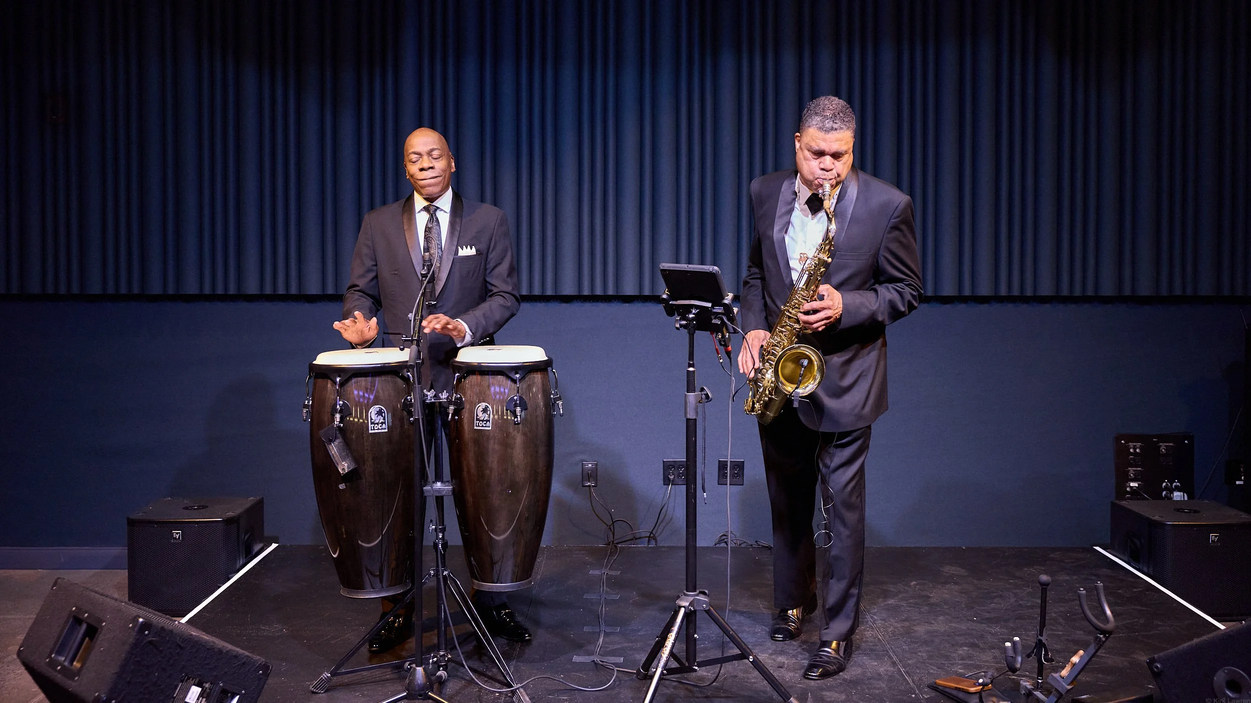 Two musicians performing on stage: one playing conga drums and the other playing a saxophone, against a dark curtain background.