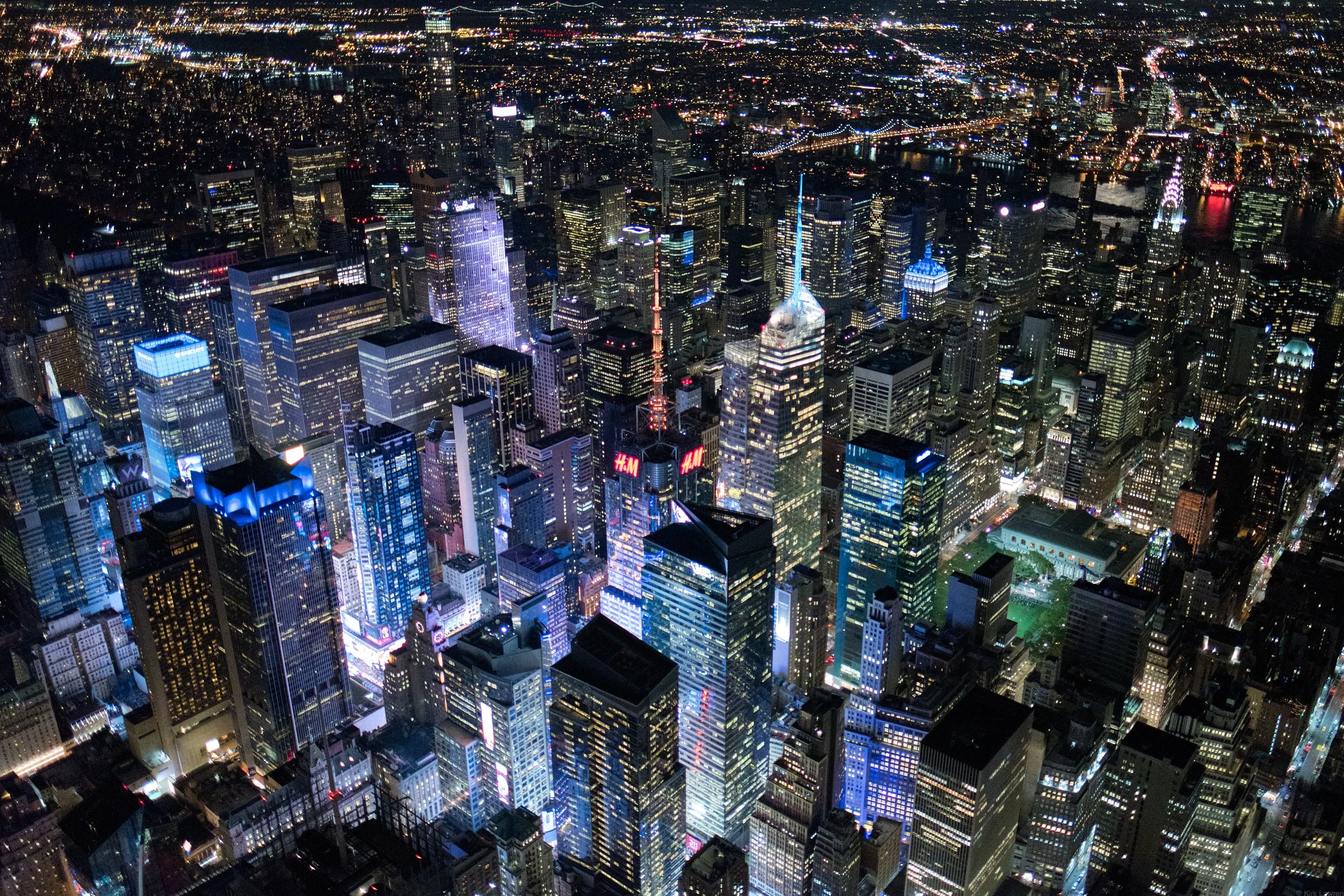 Nighttime aerial view of New York City skyline filled with illuminated skyscrapers, brightly lit streets, and bridges across the water.