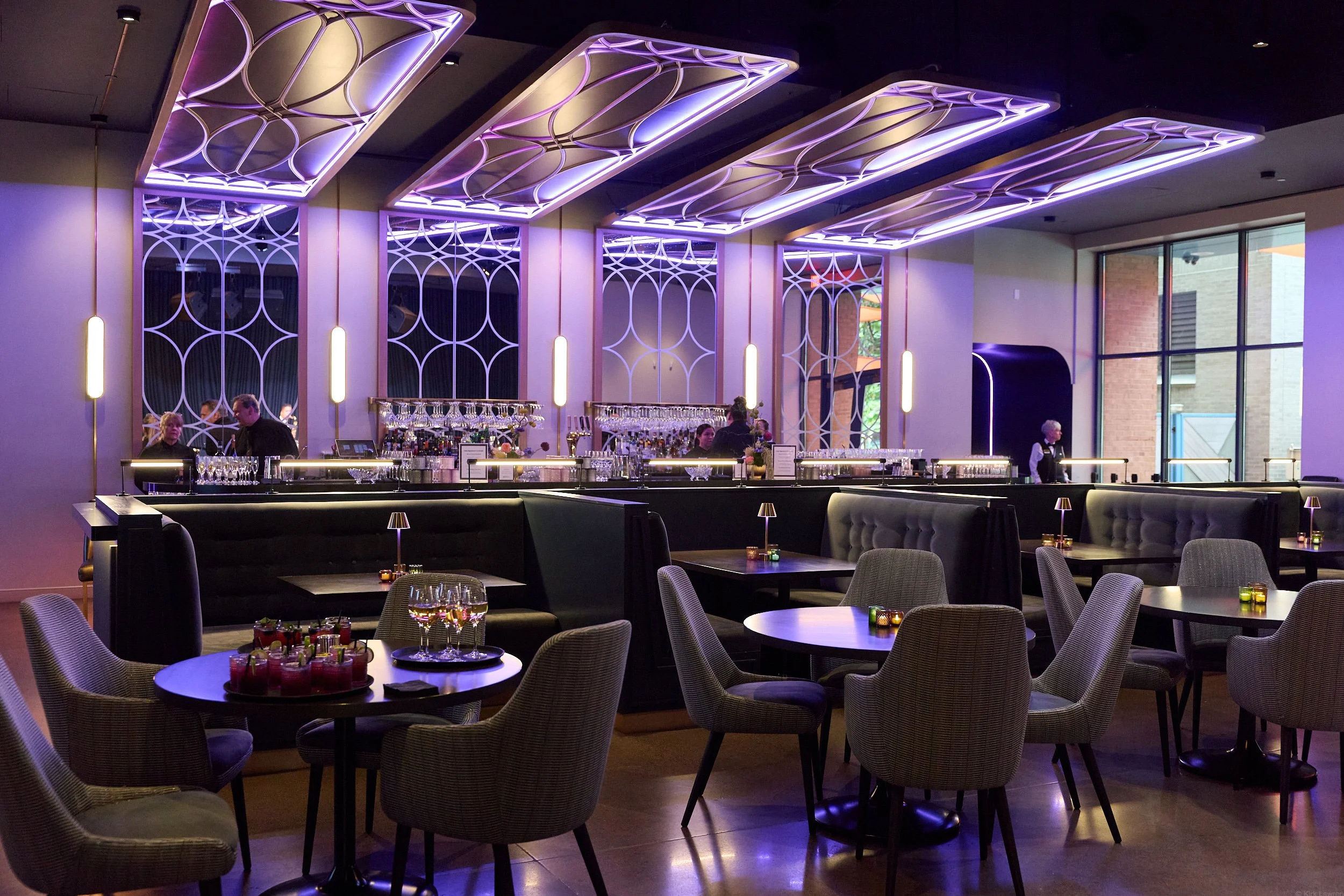 A modern interior of a bar or restaurant with purple ambient lighting. There are tables with chairs, glasses, and small candles. The bar area has a sleek counter, glassware, and a few staff members working behind it. Large windows reveal a brick buil