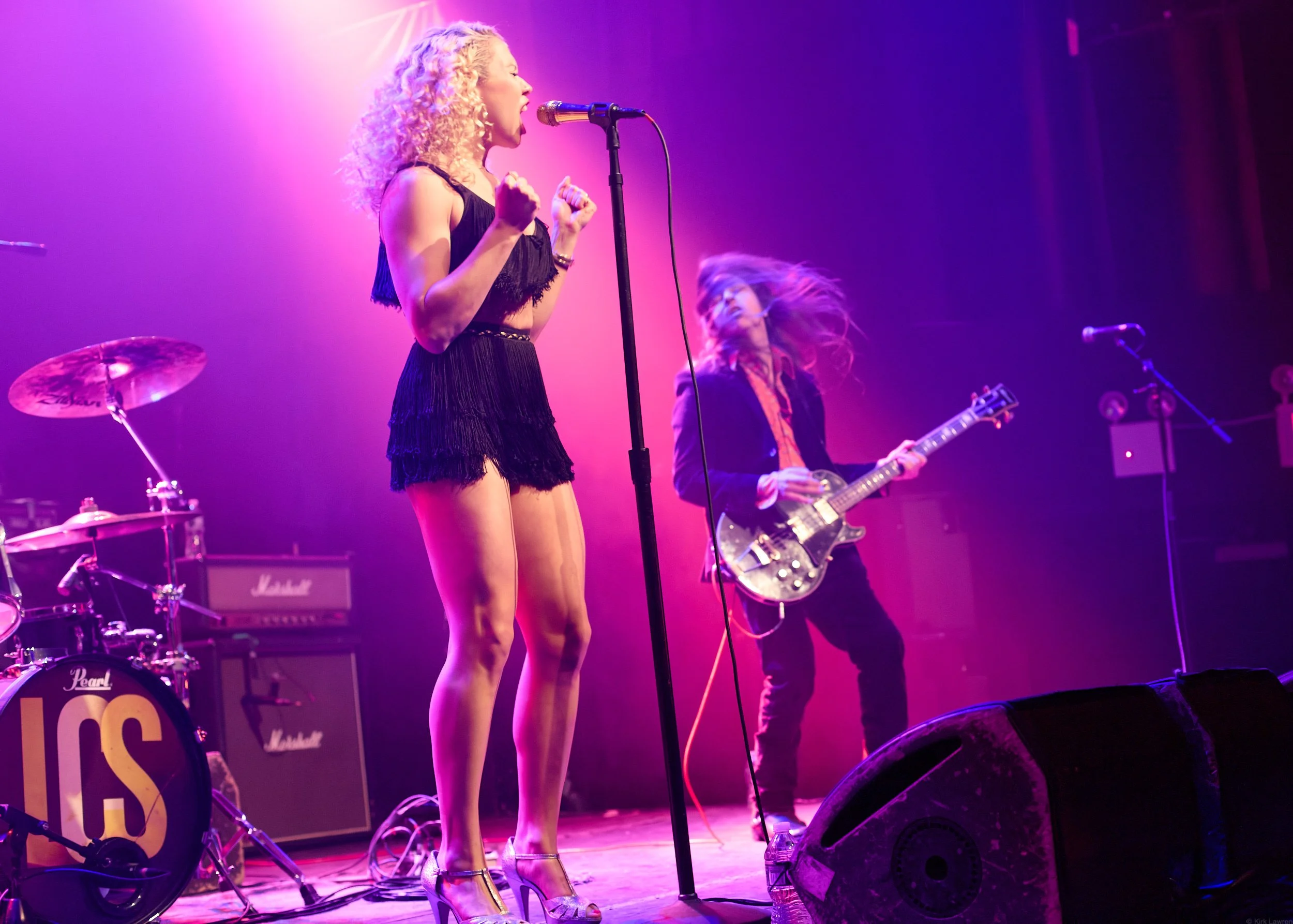 A female singer with curly blonde hair performing on stage, wearing a black dress with fringe and high heels, with a drummer and guitarist in the background under colorful stage lighting.