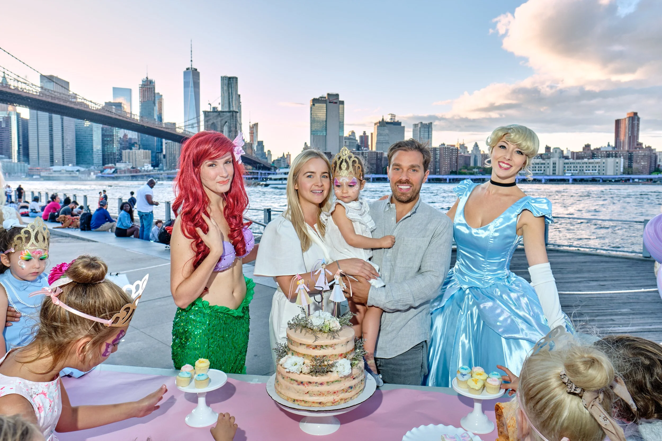 A group of children and adults celebrating a birthday outdoors on a city waterfront. The scene includes a layered birthday cake with flowers, cupcakes, and birthday decorations, with the NYC skyline and Brooklyn Bridge in the background.
