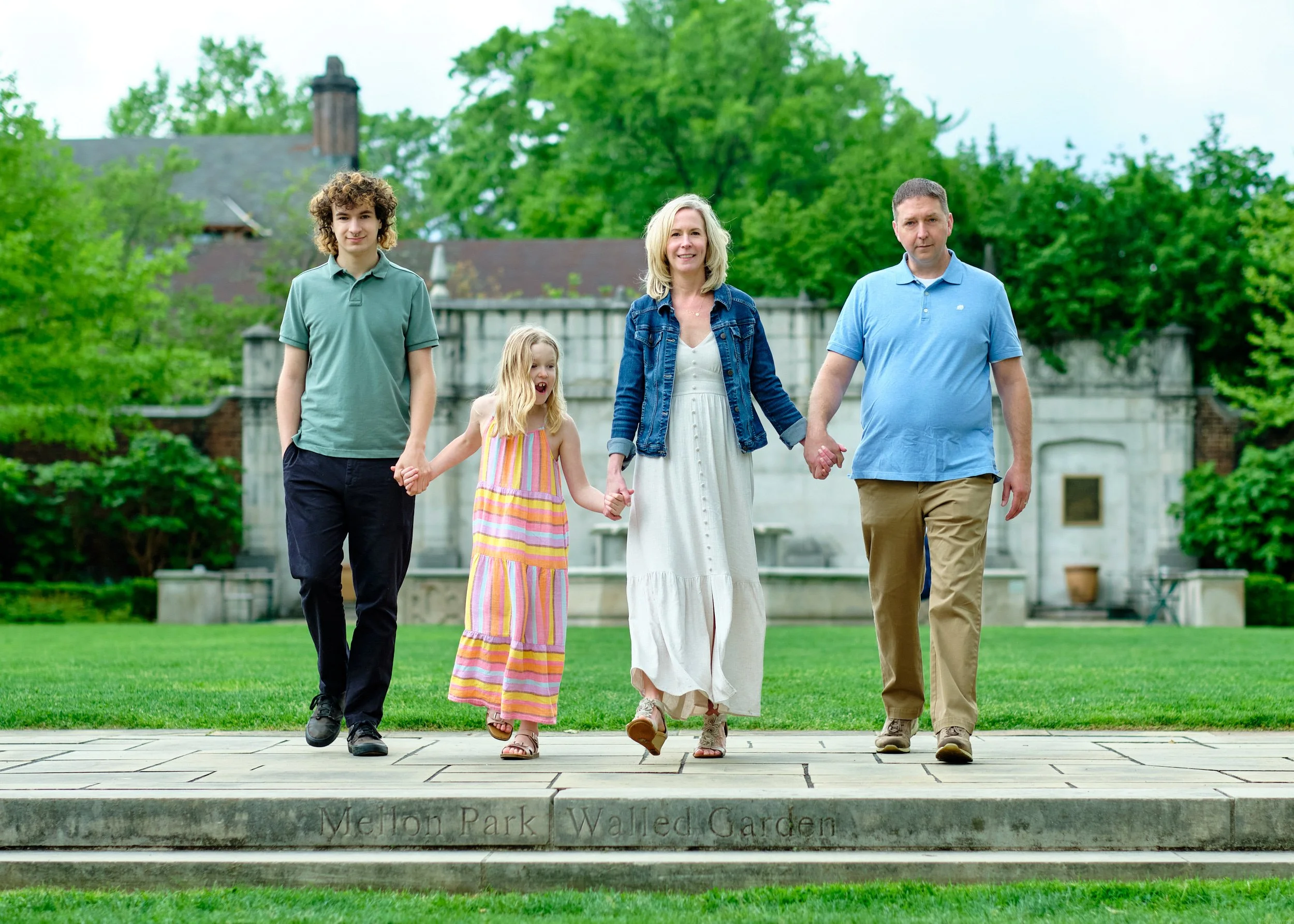 A family of five walking together outdoors in a park, holding hands and smiling. The background includes green trees and a historic-looking building.