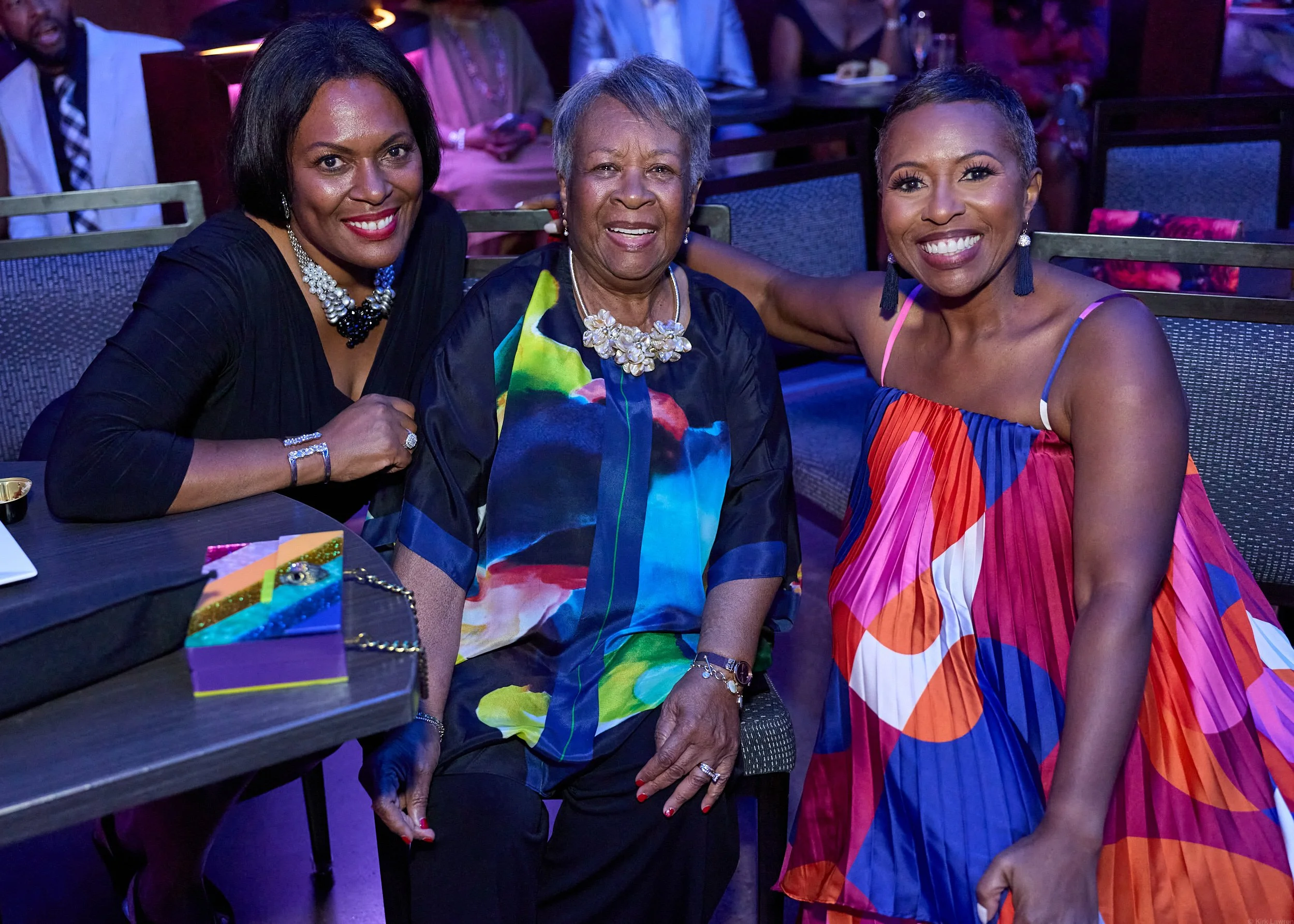 Three women sitting together at an event, smiling at the camera. They are dressed in colorful, elegant outfits and wearing jewelry. The background shows other people and purple lighting.