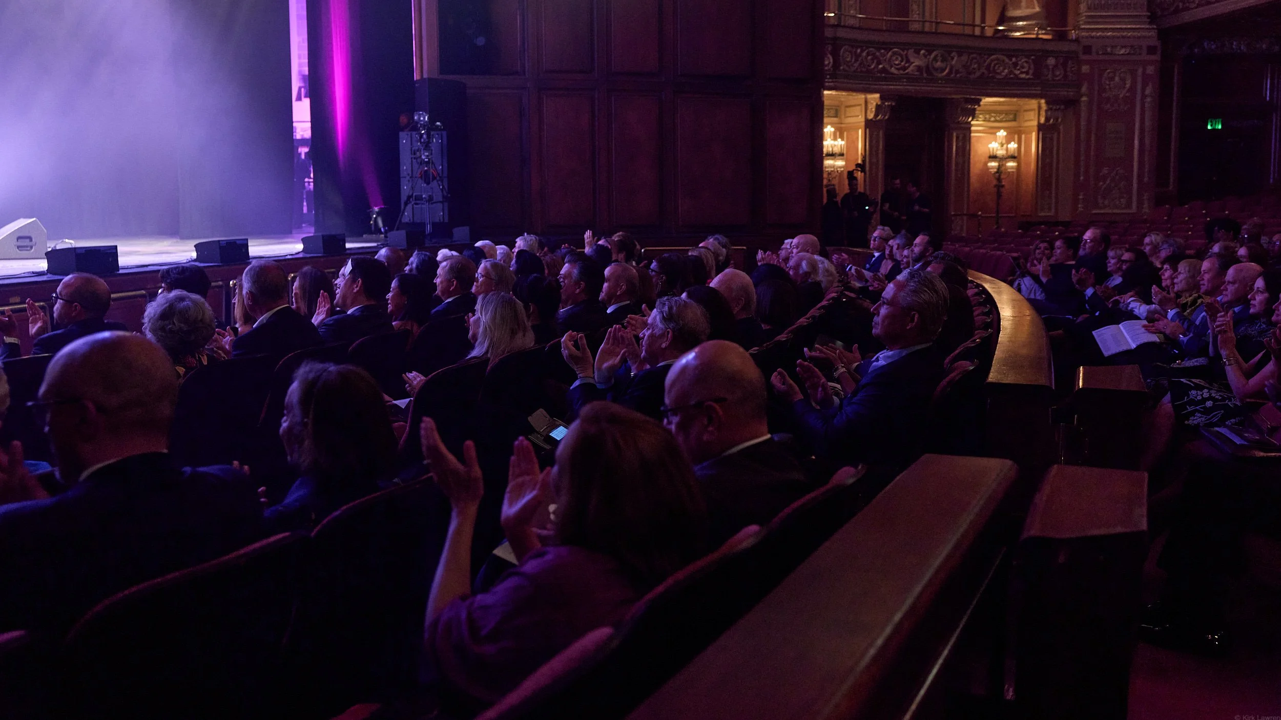 An audience in a theater watching a performance or presentation, with some clapping.
