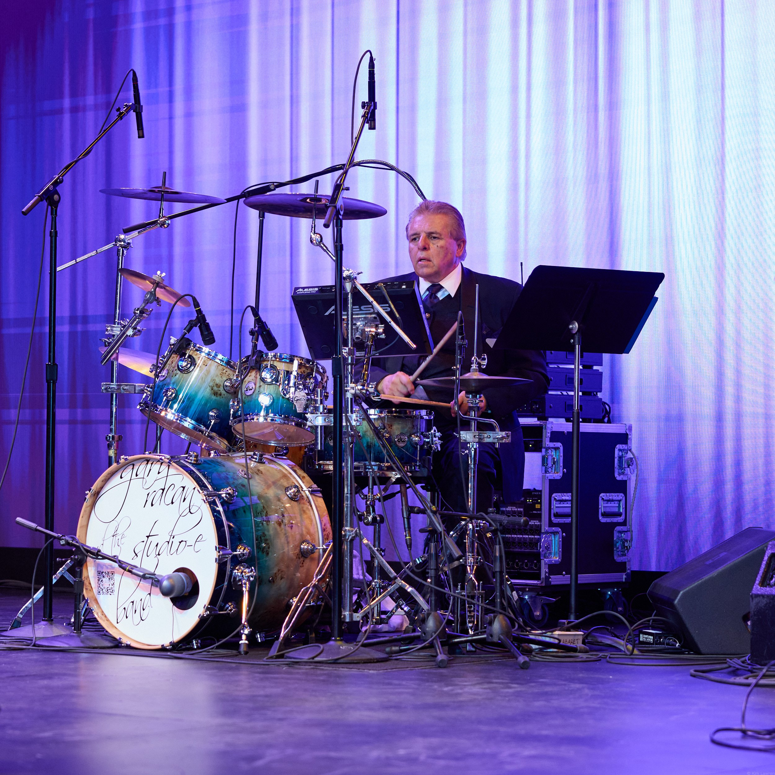 A stage with musical instruments including drums and music stands, set against a blue-purple curtain backdrop, with multiple microphones hanging above the drums.