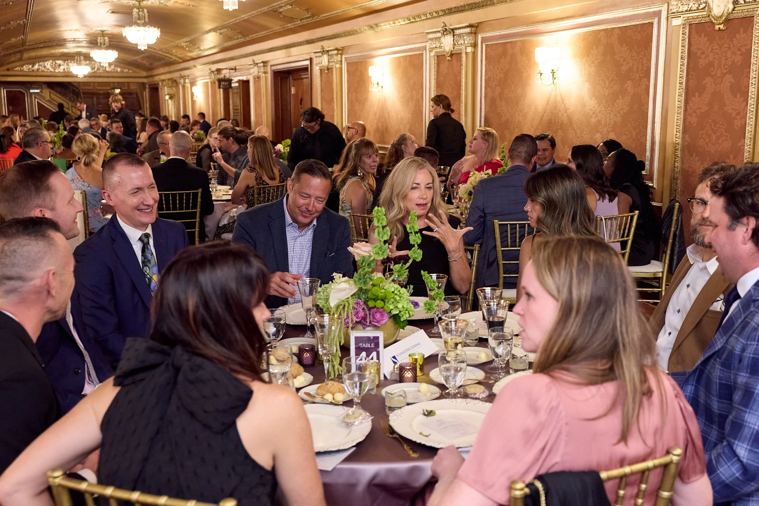 People sitting around a banquet table at a formal event in an ornate, gold-accented room with chandeliers.
