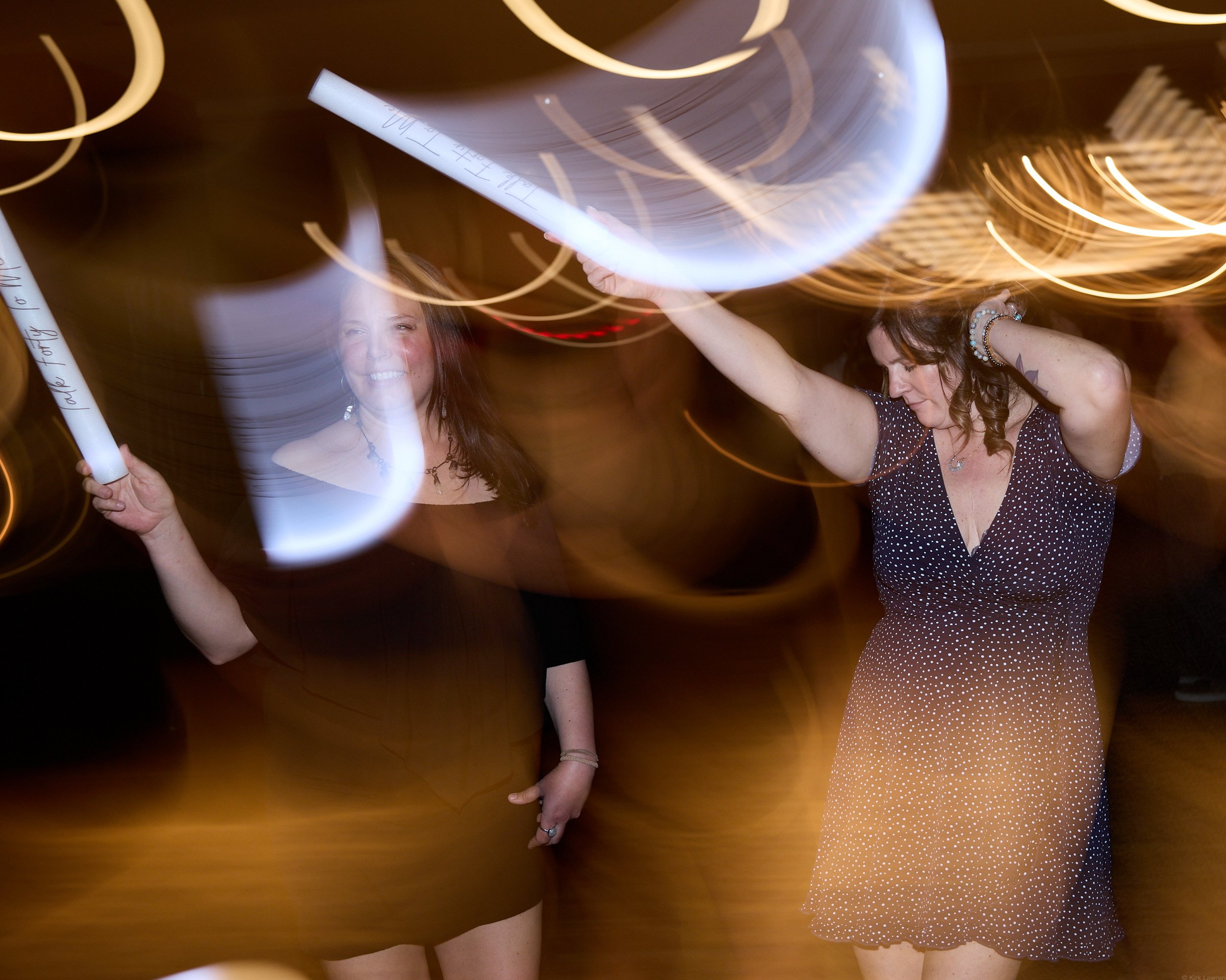 Two women dance in a dimly lit space with swirling light streaks in the background, one holding a white glow stick.