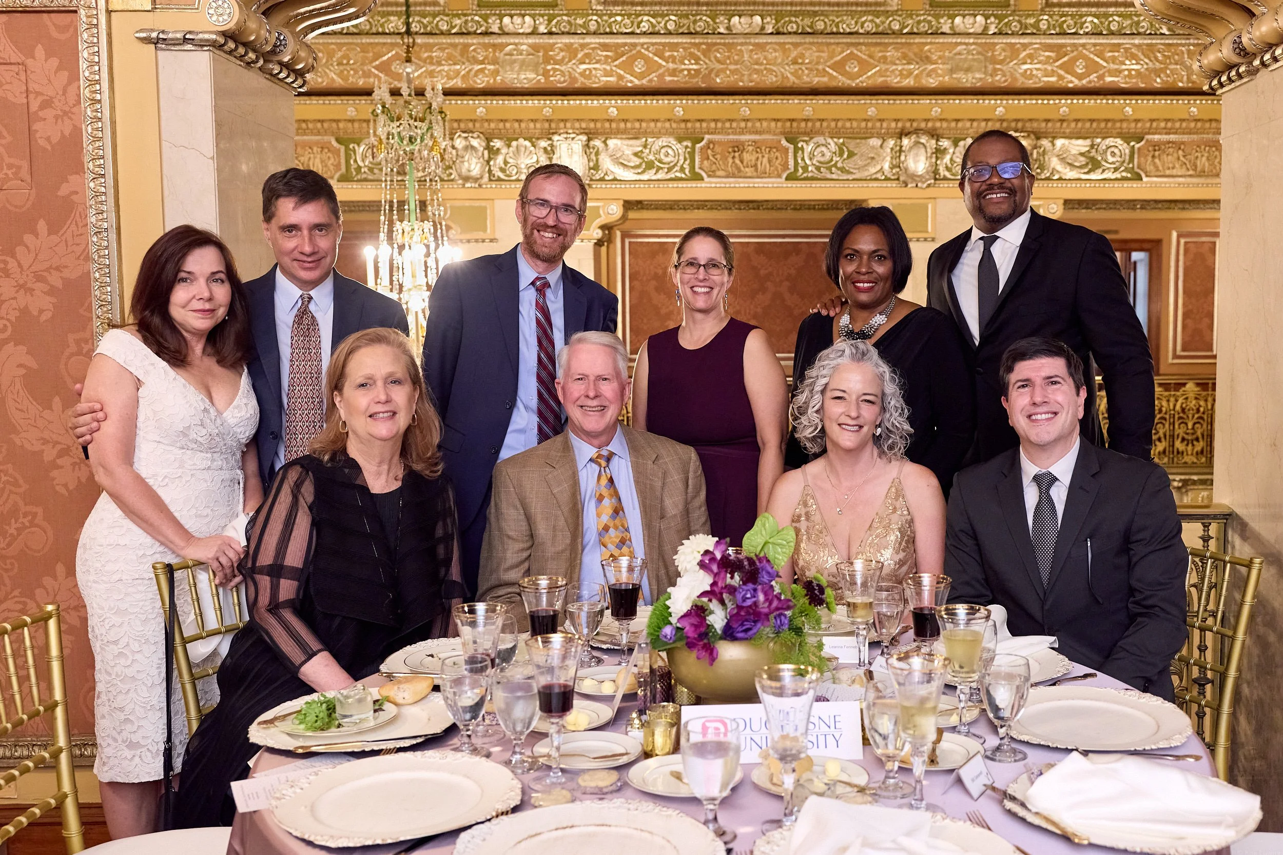 A diverse group of ten people, including men and women, posing together at a formal event or banquet in an elegant, ornately decorated room with gold accents, chandeliers, and floral wallpaper. They are dressed in formal attire, smiling for the camer