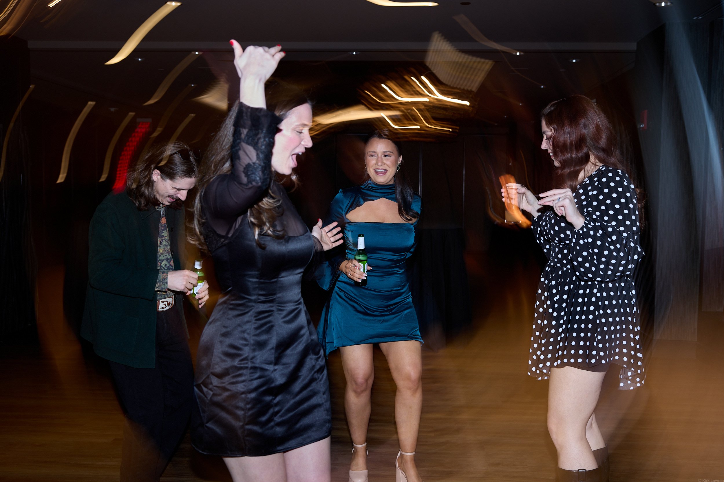 Five women dancing and socializing at a party in a dimly lit indoor venue, holding drinks.