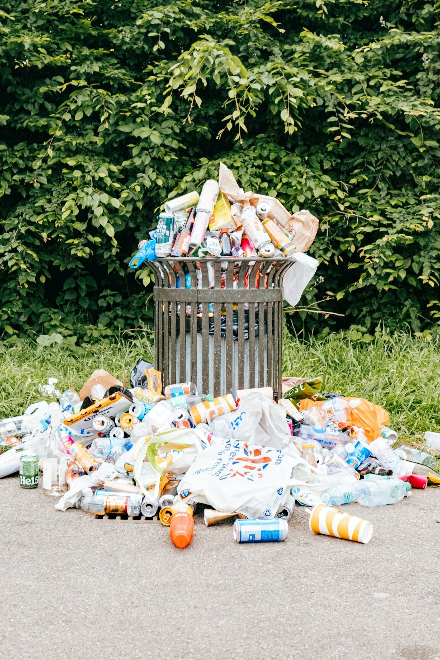 Overflowing trash can with scattered plastic bottles, cans, and paper on the ground in front, set against a background of green bushes.