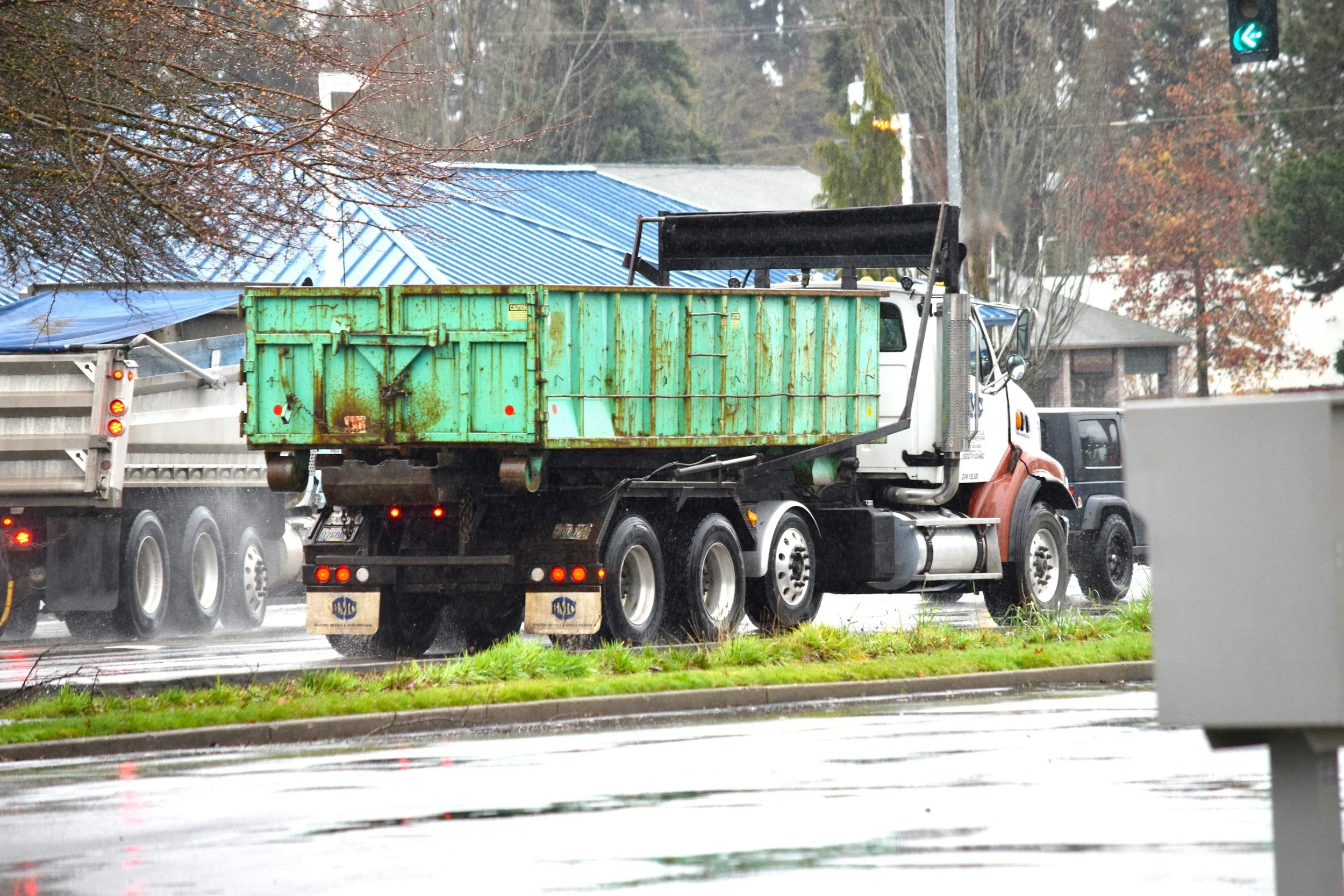 A semi-truck with an orange cab and green cargo container driving on a wet road during rain, with a traffic light showing a green arrow, trees without leaves, and a house with a blue roof in the background.