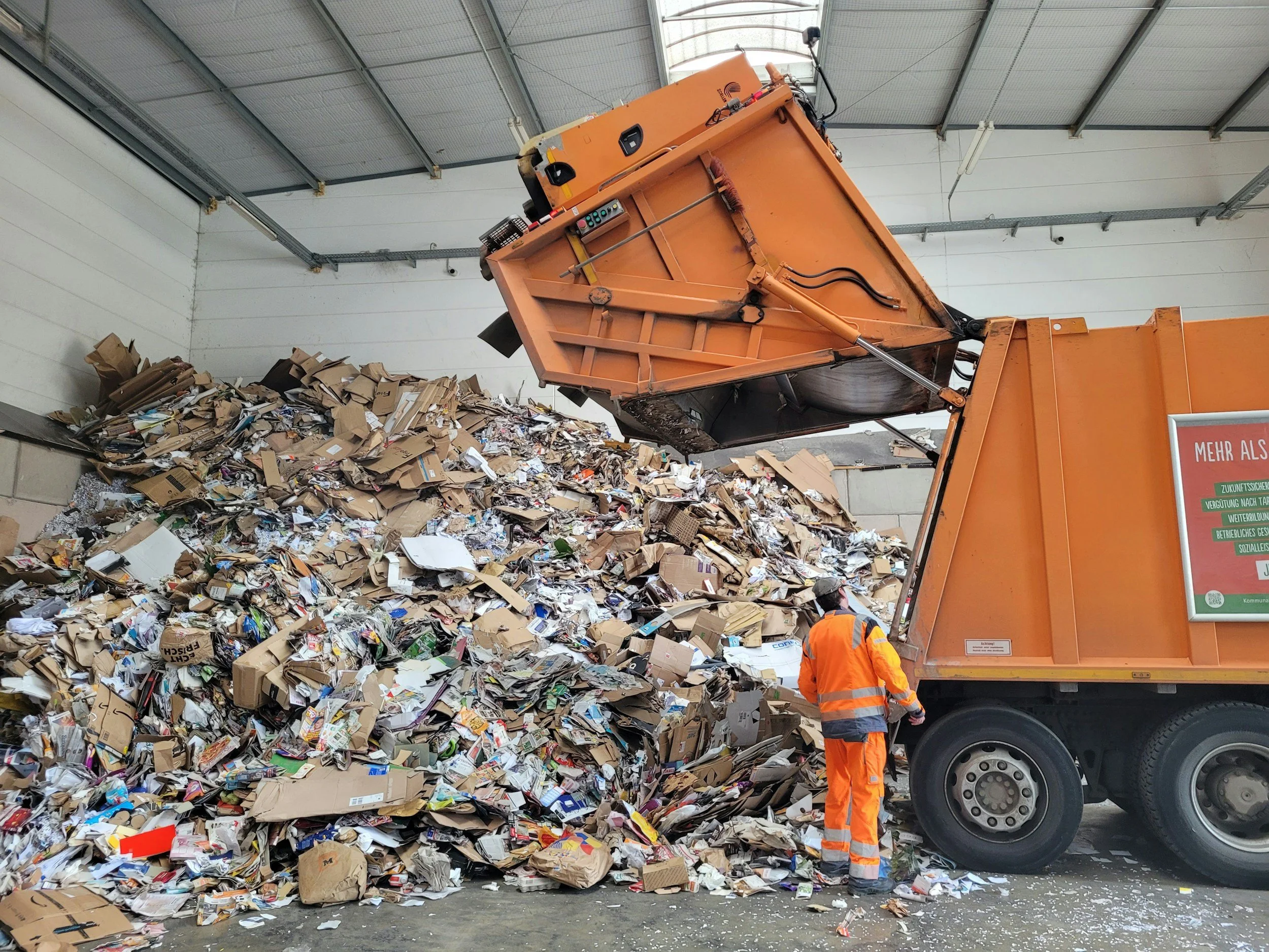 A large orange garbage truck is dumping a tall pile of mixed waste and paper onto the ground inside a covered recycling facility, with two workers in orange safety uniforms observing.