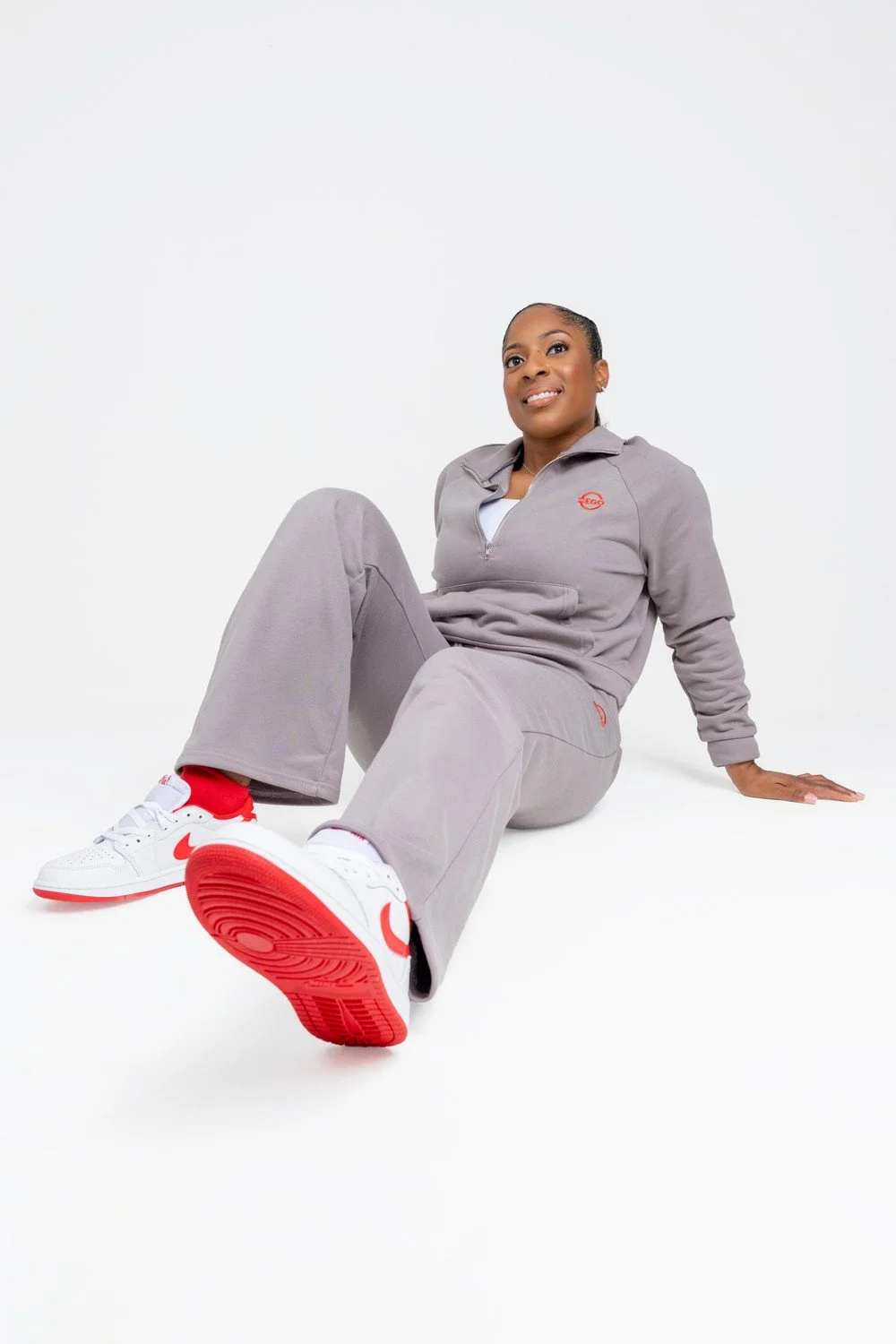 A woman sitting on the floor wearing a gray tracksuit, white and red sneakers, smiling, against a white background.
