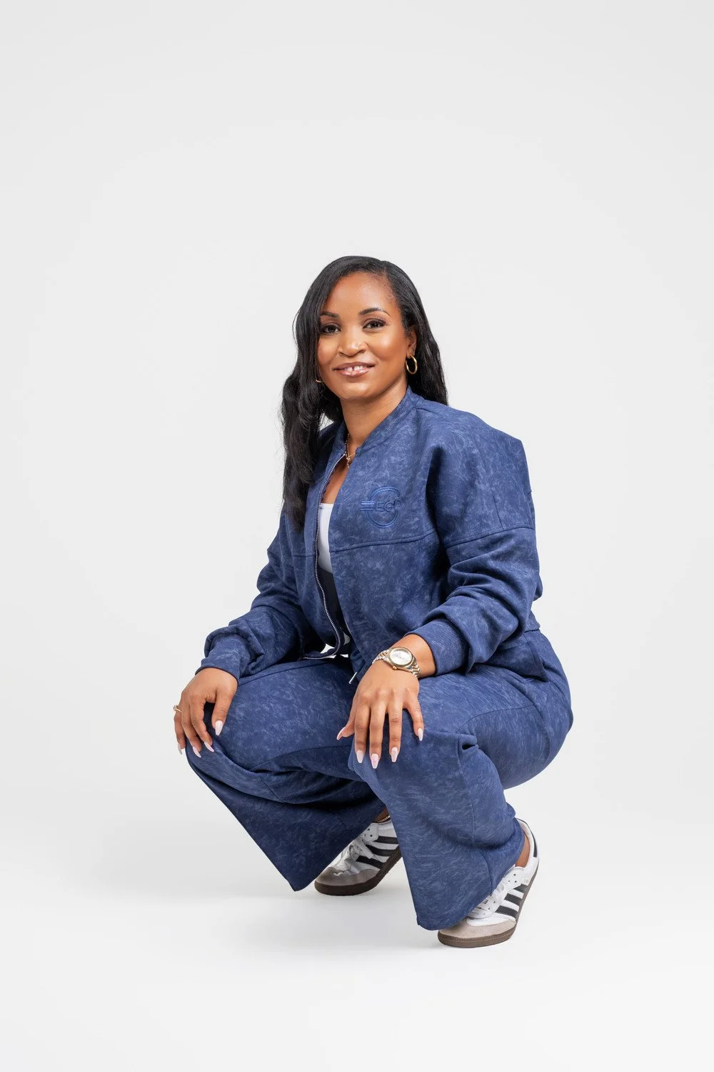 A woman in a matching blue casual outfit with a zip-up jacket and wide-leg pants, squatting and smiling at the camera, against a plain white background.