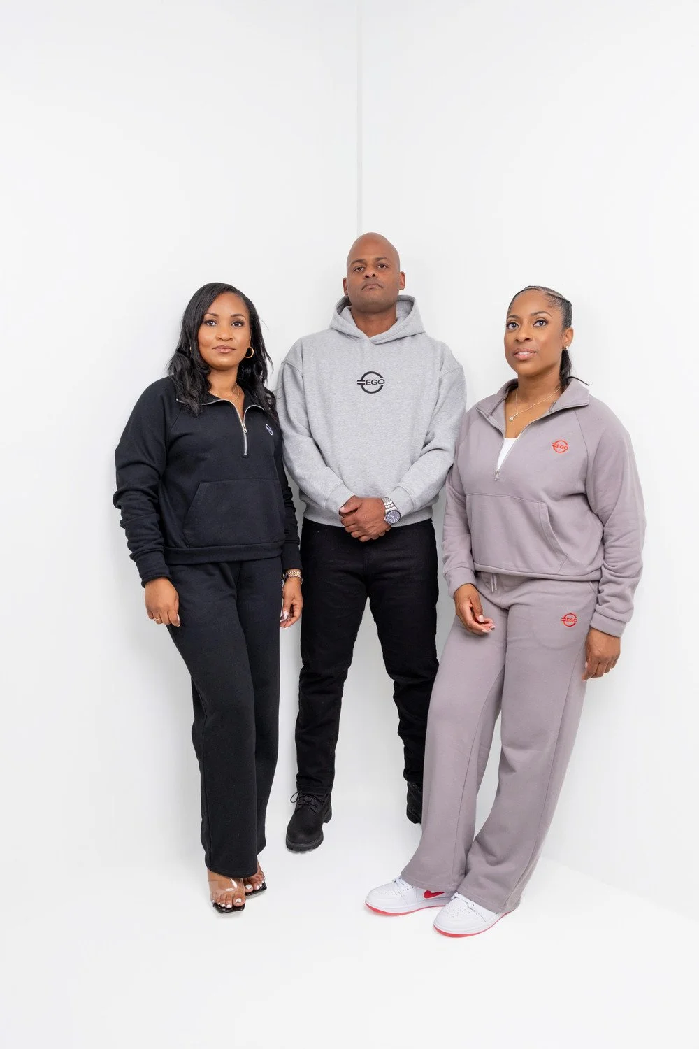 Three people standing against a plain white wall, wearing casual sportswear. From left to right: a woman in black sweatshirt and pants, a man in a gray hoodie and black pants, and a woman in beige tracksuit with white sneakers.